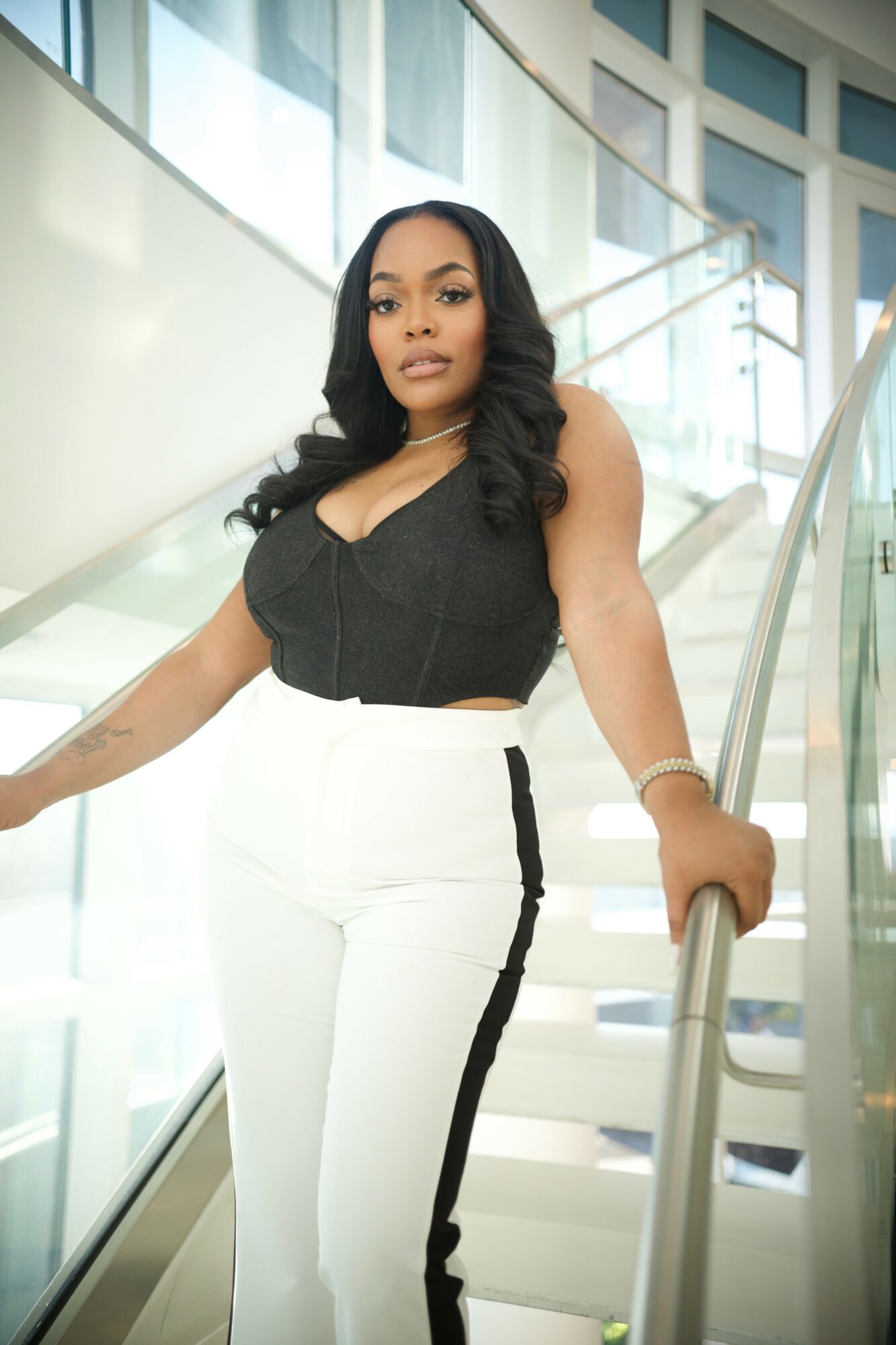 Woman with long dark hair in black top and white pants standing on a staircase with glass railing inside a modern building.