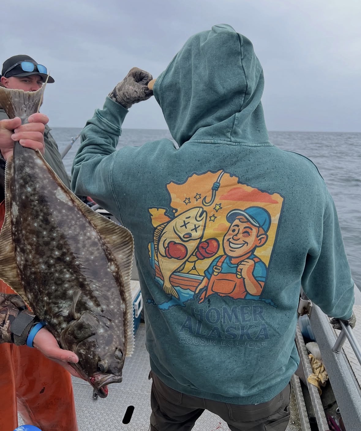 Person in teal hoodie with cartoon fish and smiling character on back, holding fishing rod on boat, ocean in background.