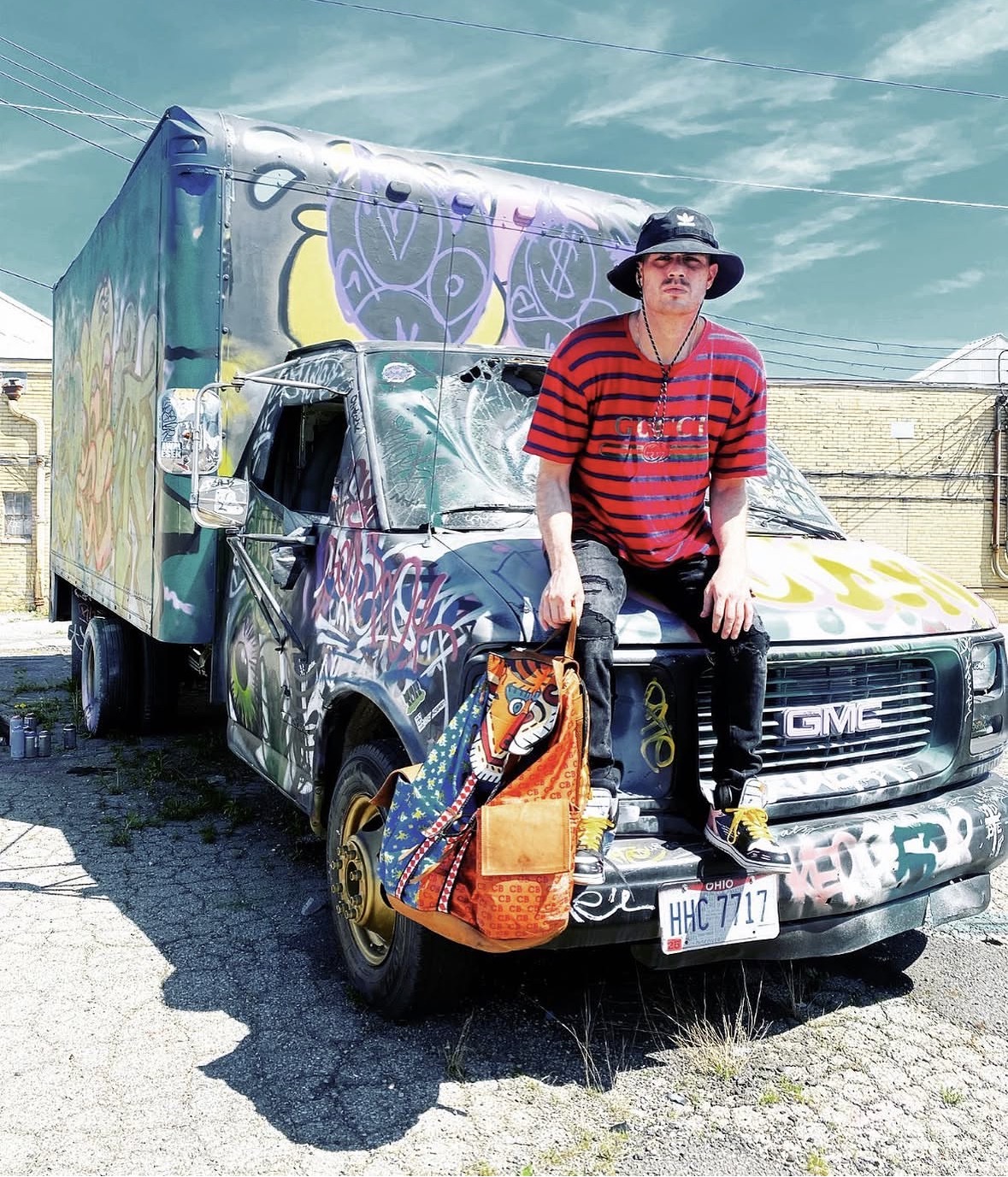 Person wearing a hat and sunglasses, standing on a graffiti-covered GMC truck with a colorful backpack, outdoors under a blue sky.