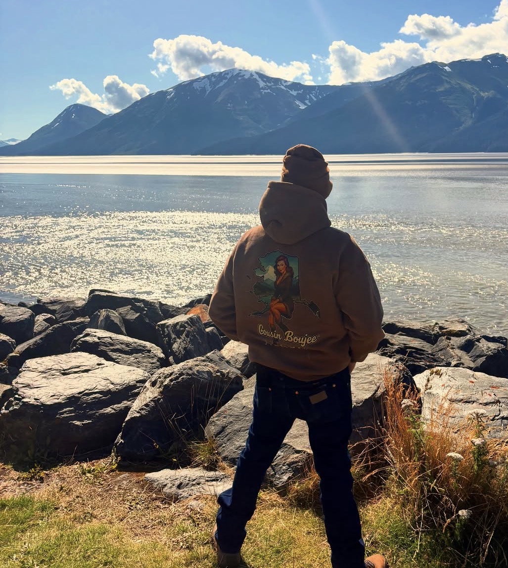 Person standing on rocky shore facing a lake with mountains and sky in background.