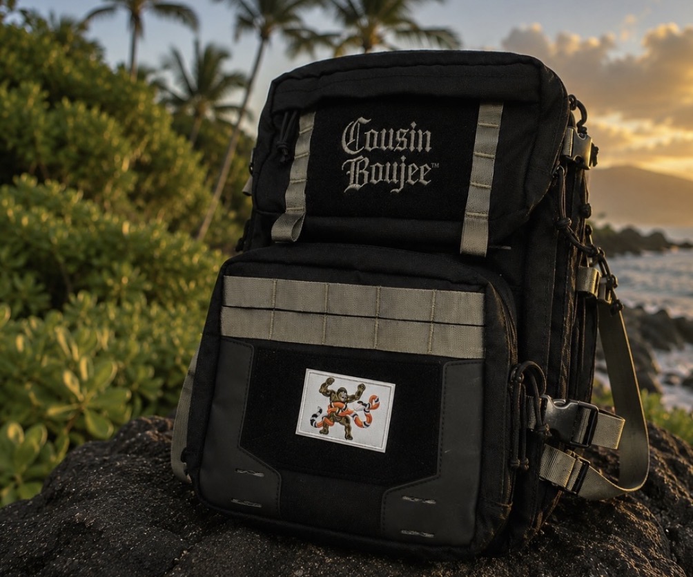 Black backpack with gray accents and a gingerbread man sticker, outdoors near water and greenery, sunset background.