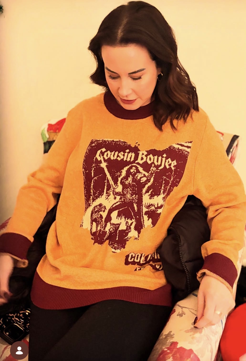 Woman with dark hair wearing a yellow sweater with a graphic design, sitting on a chair, looking down.