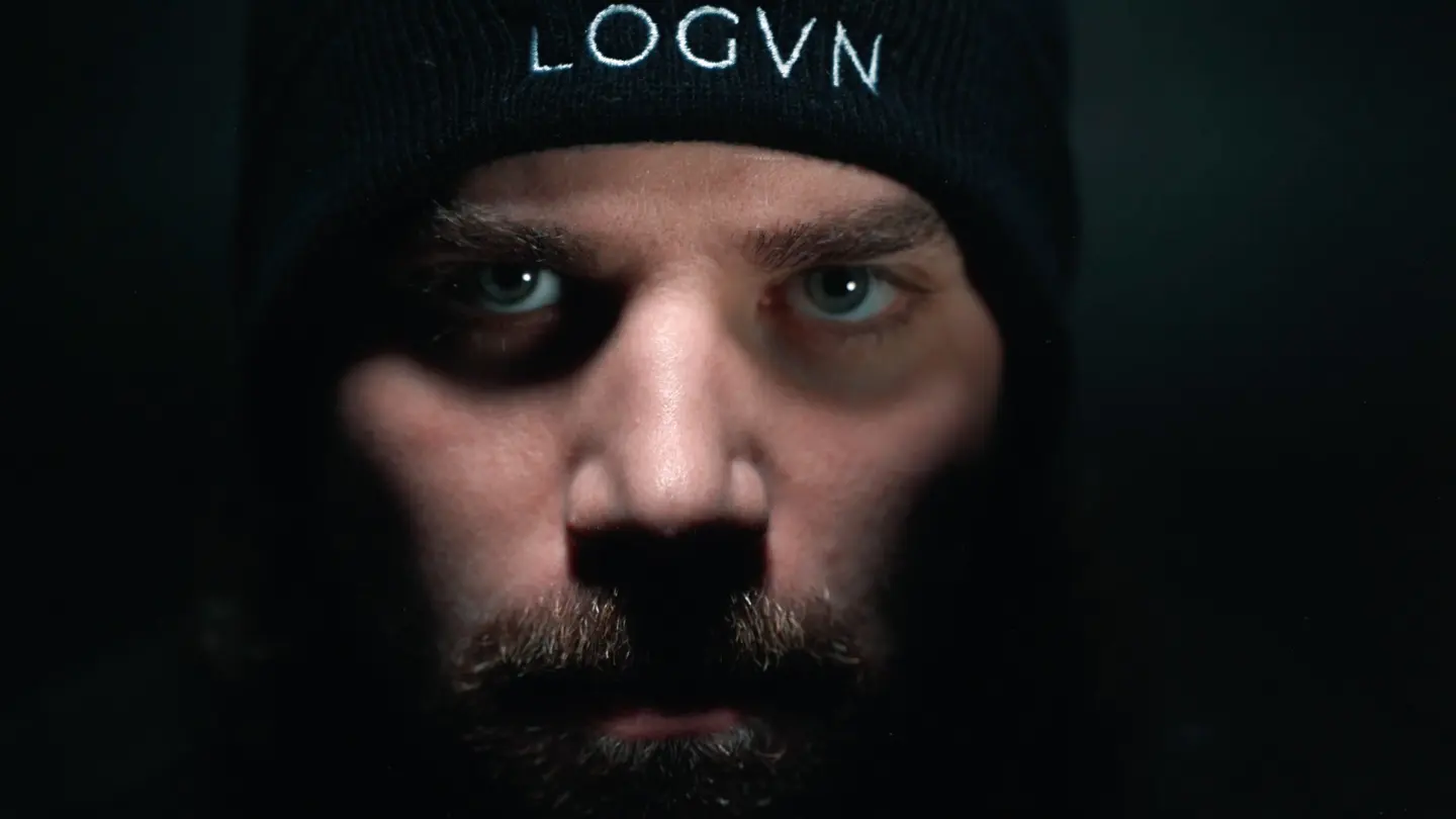 Close-up of a person's face with intense expression, wearing a black beanie with white text, dark background.