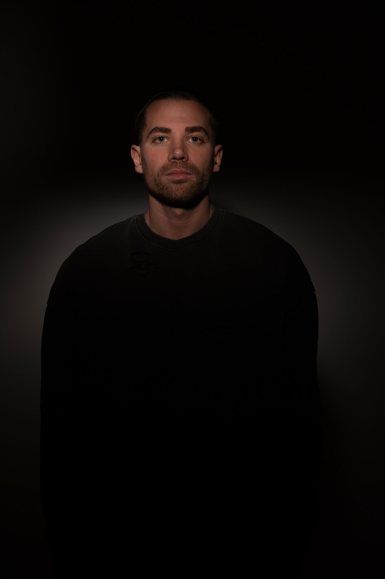 Portrait of a man with short dark hair and beard, wearing a dark shirt, against a dark background.