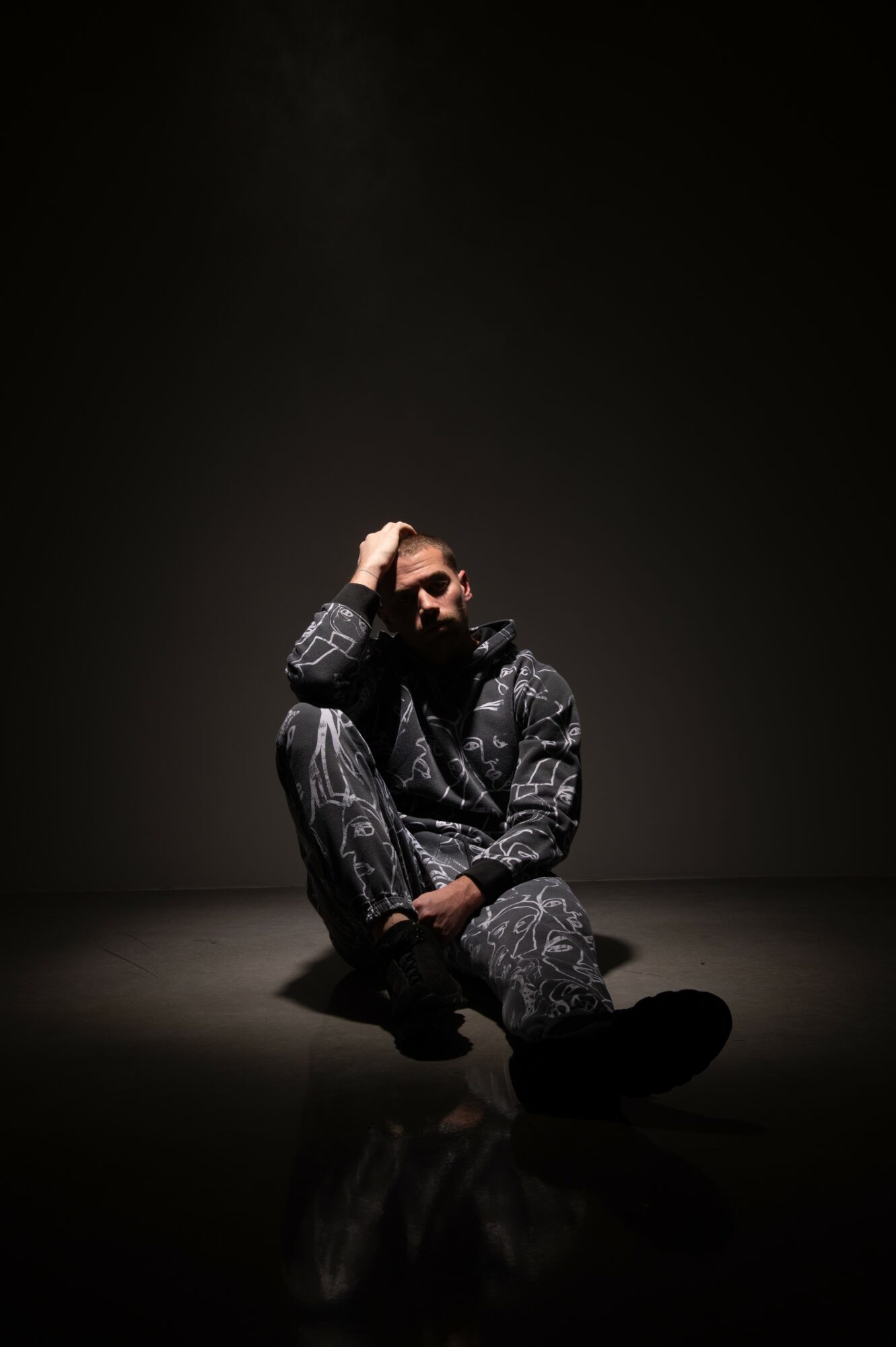Person sitting on the floor in a dark setting, wearing patterned clothing, with one hand on their head.