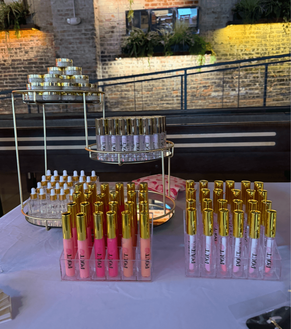 Display of pink and clear bottles with gold caps on a table, with a brick wall and outdoor scene in background.