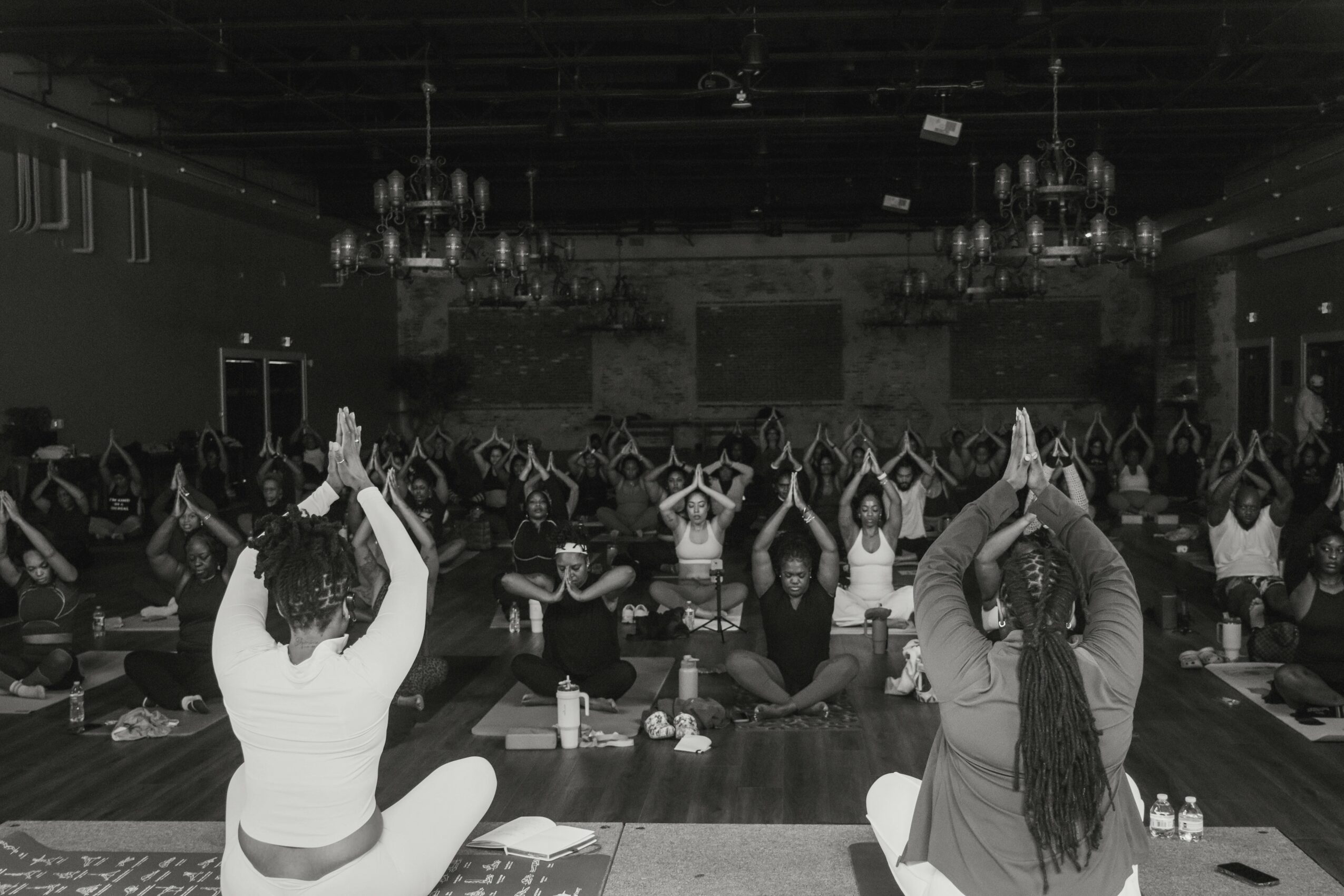 Group of people practicing yoga in a large indoor space, sitting on mats with hands raised overhead, facing forward.