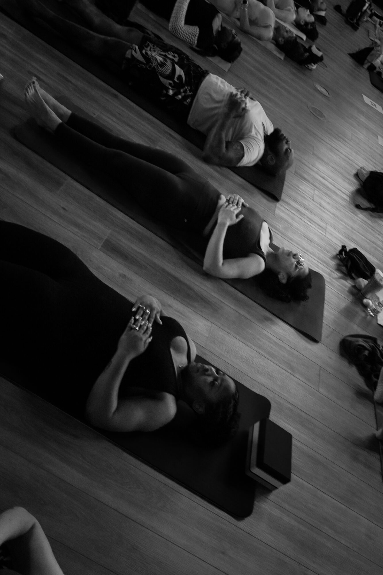 People lying on yoga mats in a room, some holding drinks, with wooden flooring and bags nearby.