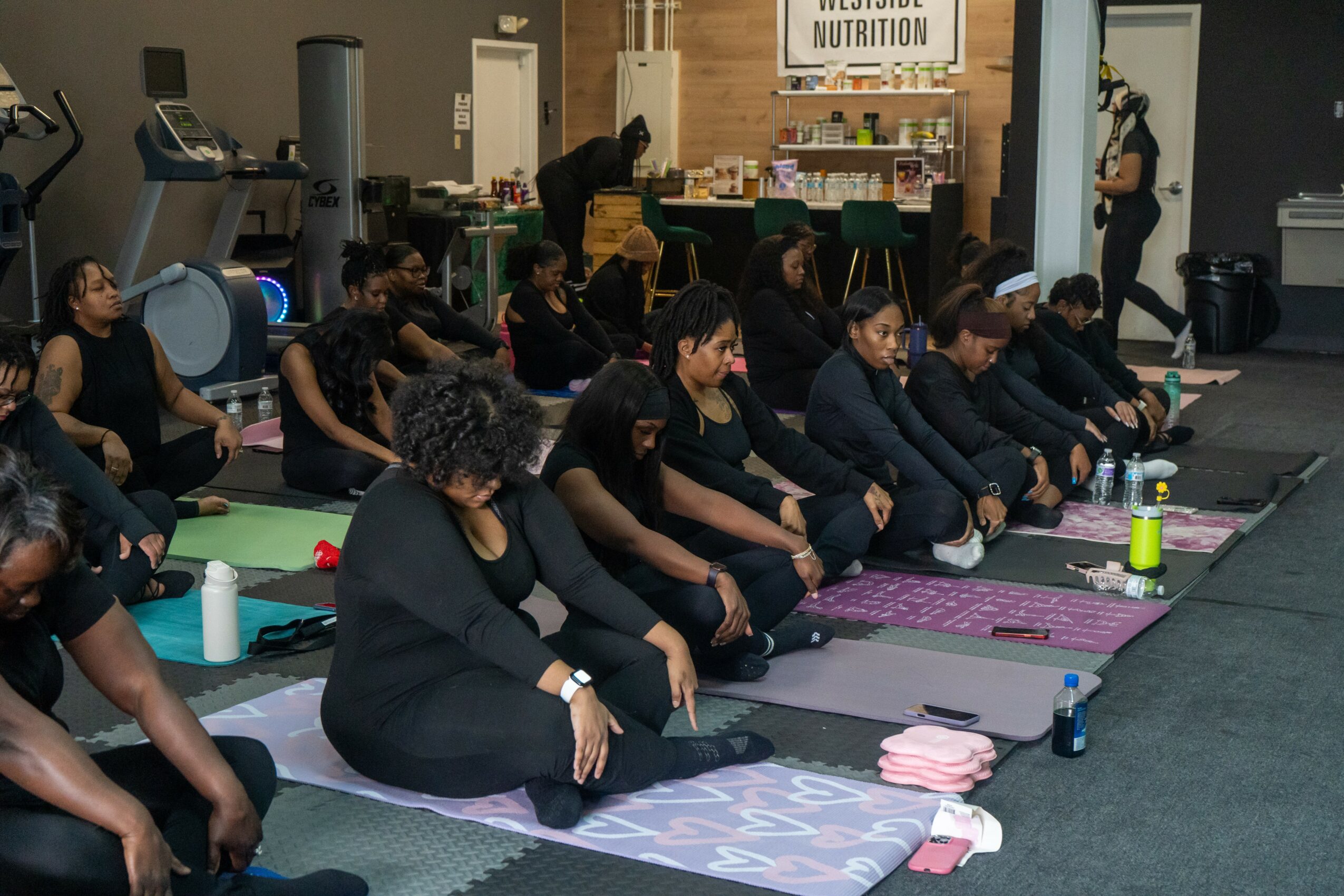 Group of people sitting cross-legged on mats in a fitness class, facing forward, in a gym or studio setting.