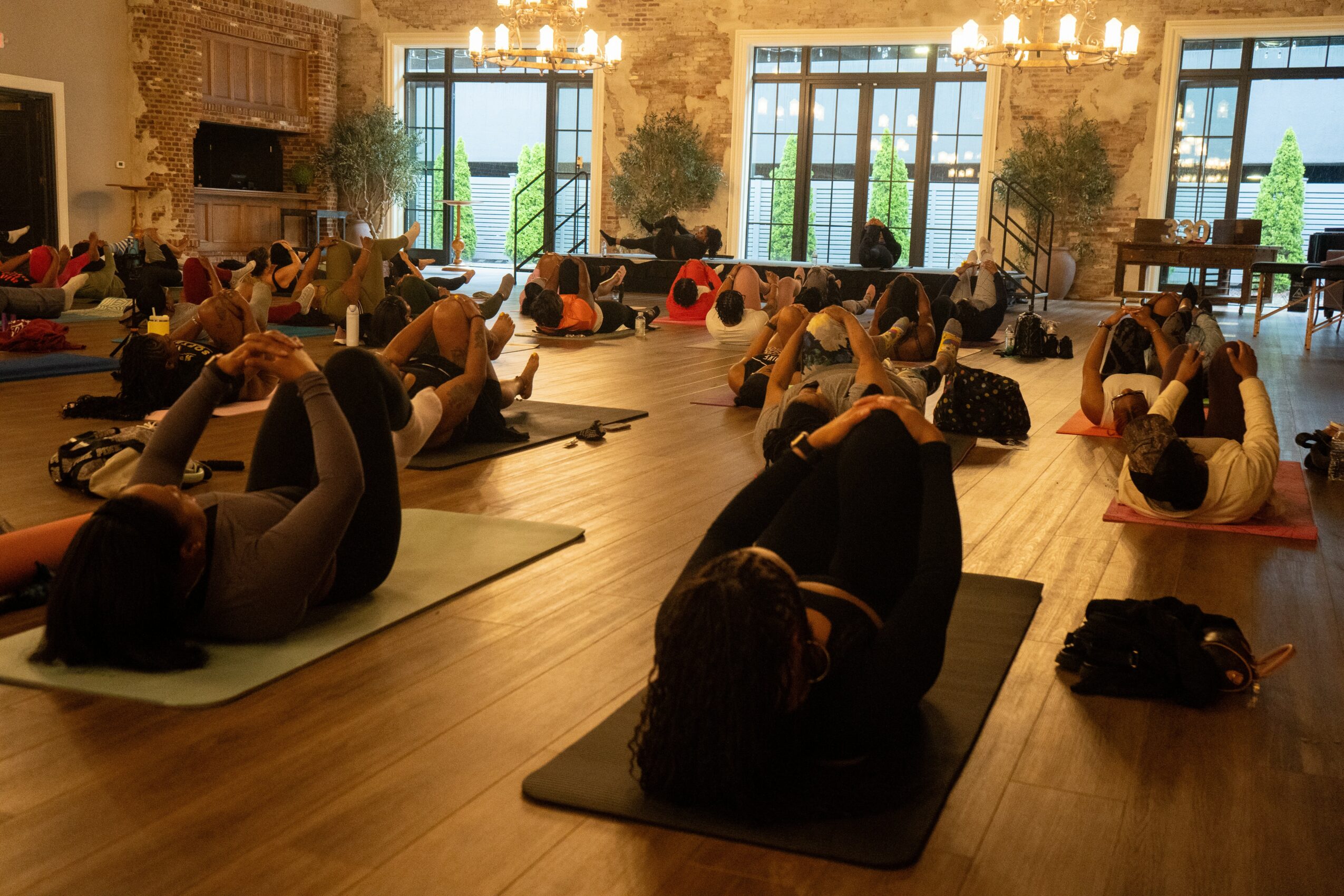 People participating in a group yoga or stretching class in a spacious, well-lit room with large windows and wooden floors.