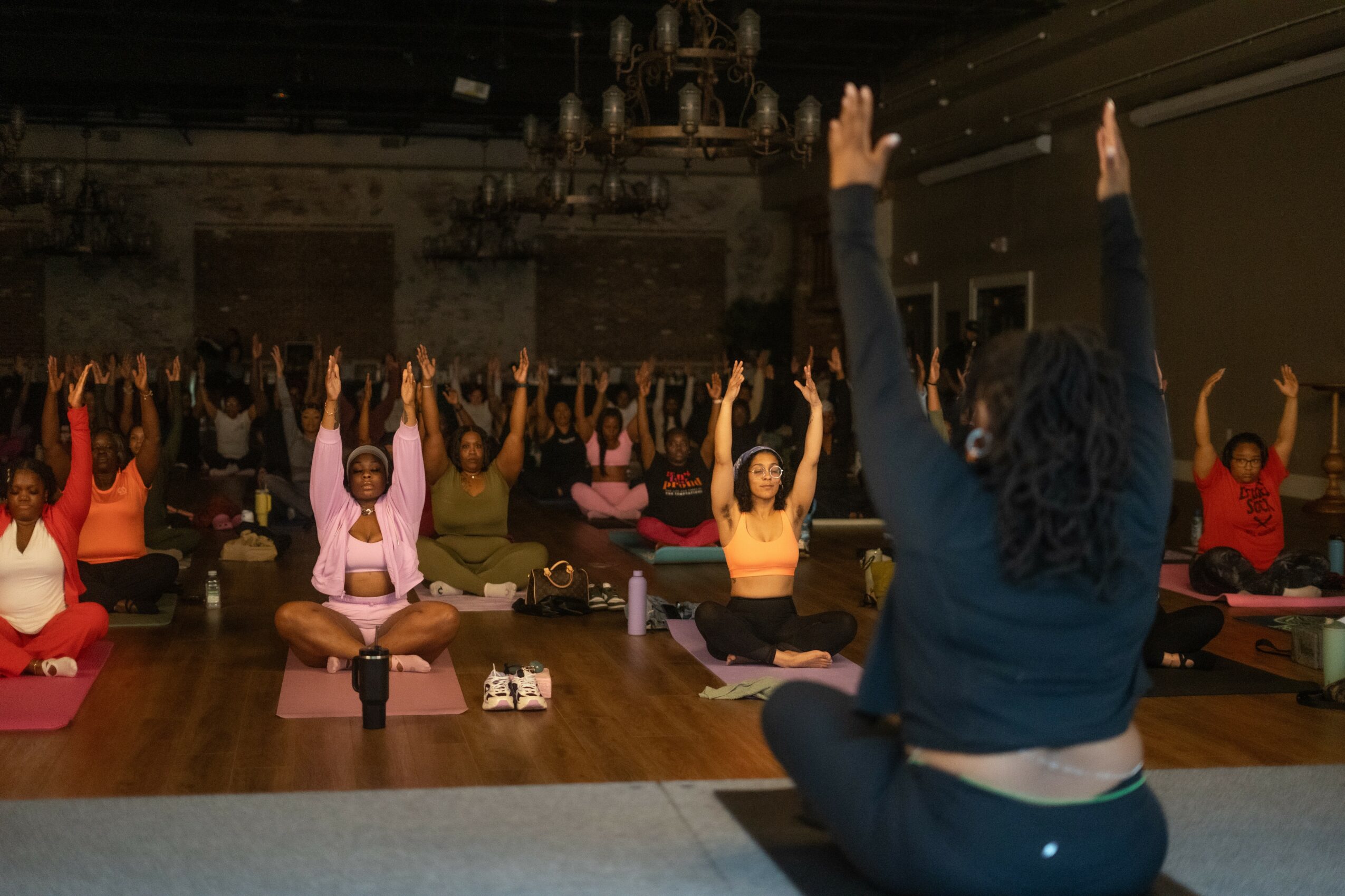 Group of people practicing yoga in a dimly lit room, sitting cross-legged with arms raised overhead, led by instructor at front.