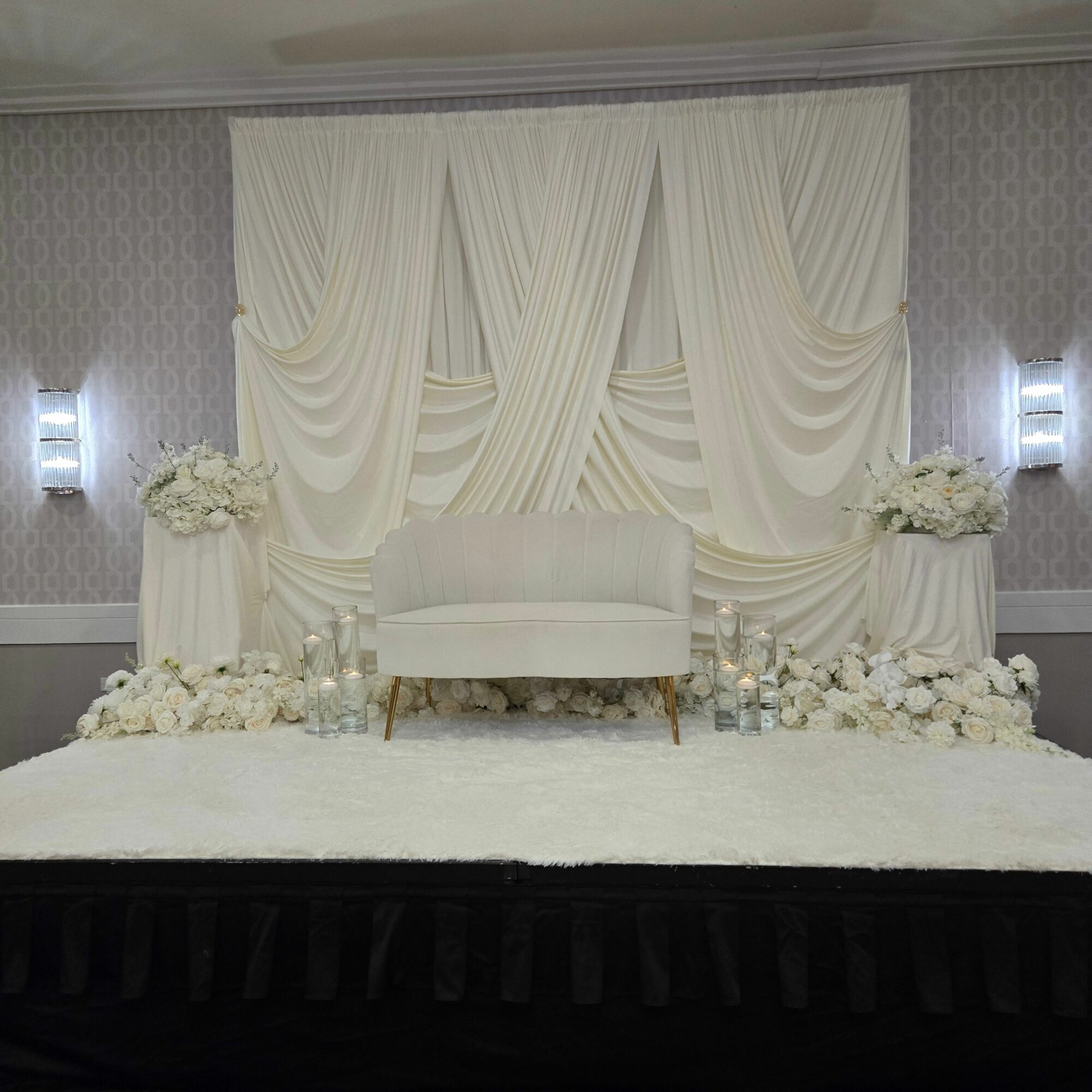 Elegant wedding stage with white drapery, floral arrangements, and soft lighting, decorated for a celebration or ceremony.