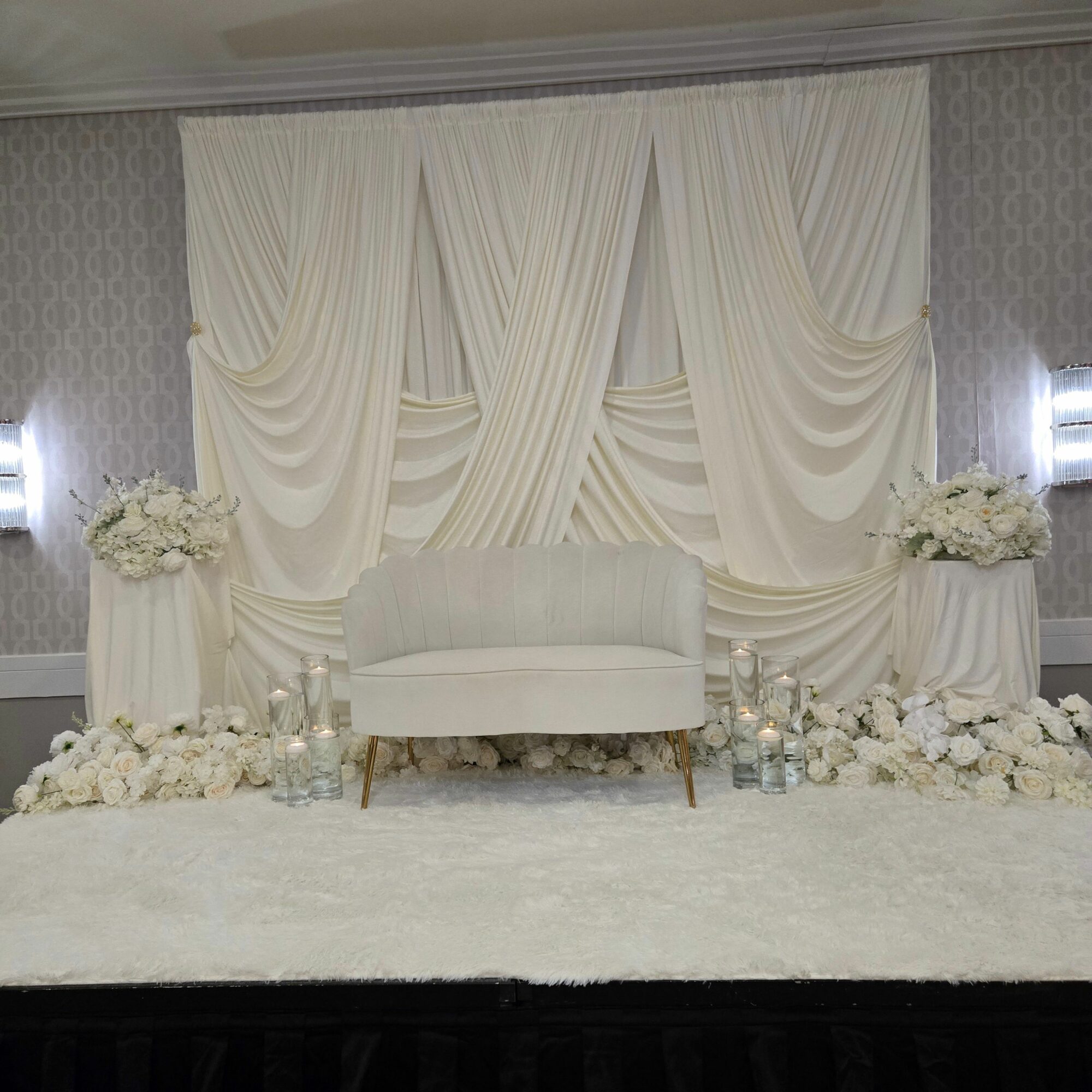 Wedding altar with white drapery, floral arrangements, and a white bench, decorated for a ceremony.