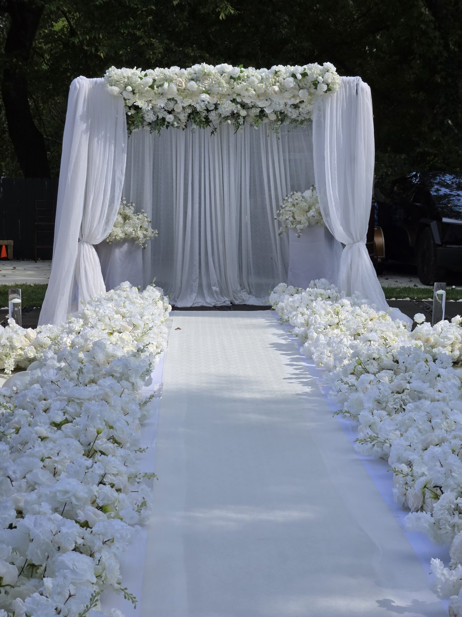 Wedding aisle with white flowers and draped fabric, leading to an arch decorated with flowers, outdoors.