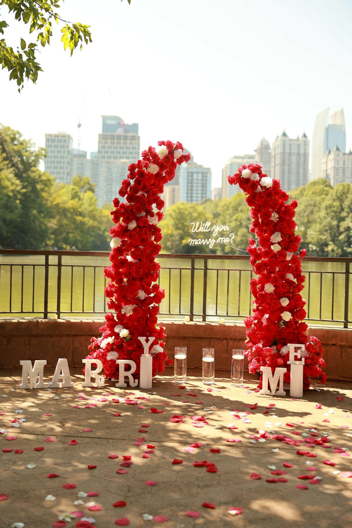 Two tall red flower arrangements on white stands with scattered petals on the ground, set outdoors with trees and city buildings in background.