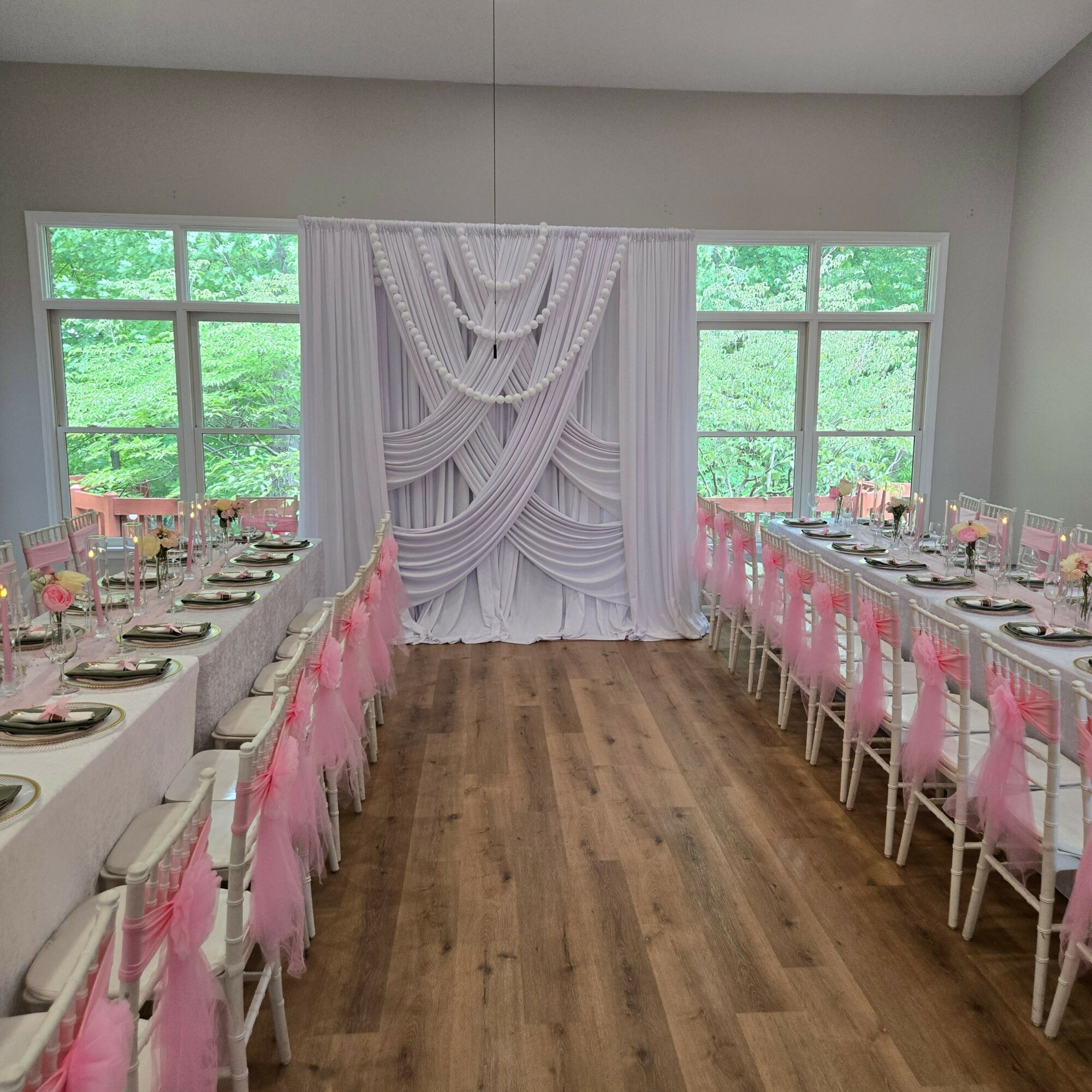 Long dining table set with plates, glasses, and pink napkins, in a bright room with large windows and wooden floor.
