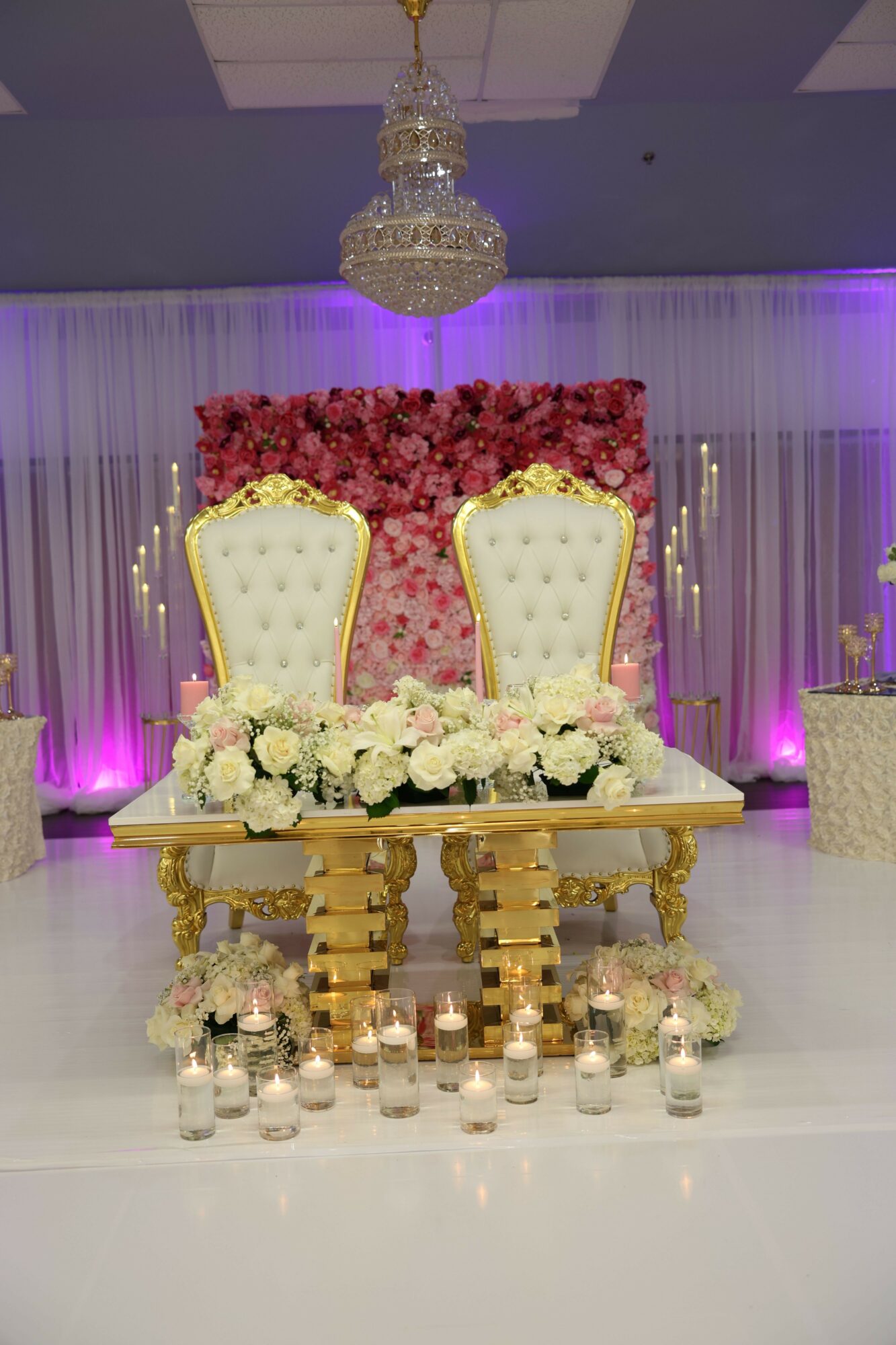 Elegant table setup with two white chairs, floral backdrop, and gold accents, illuminated by purple lighting.
