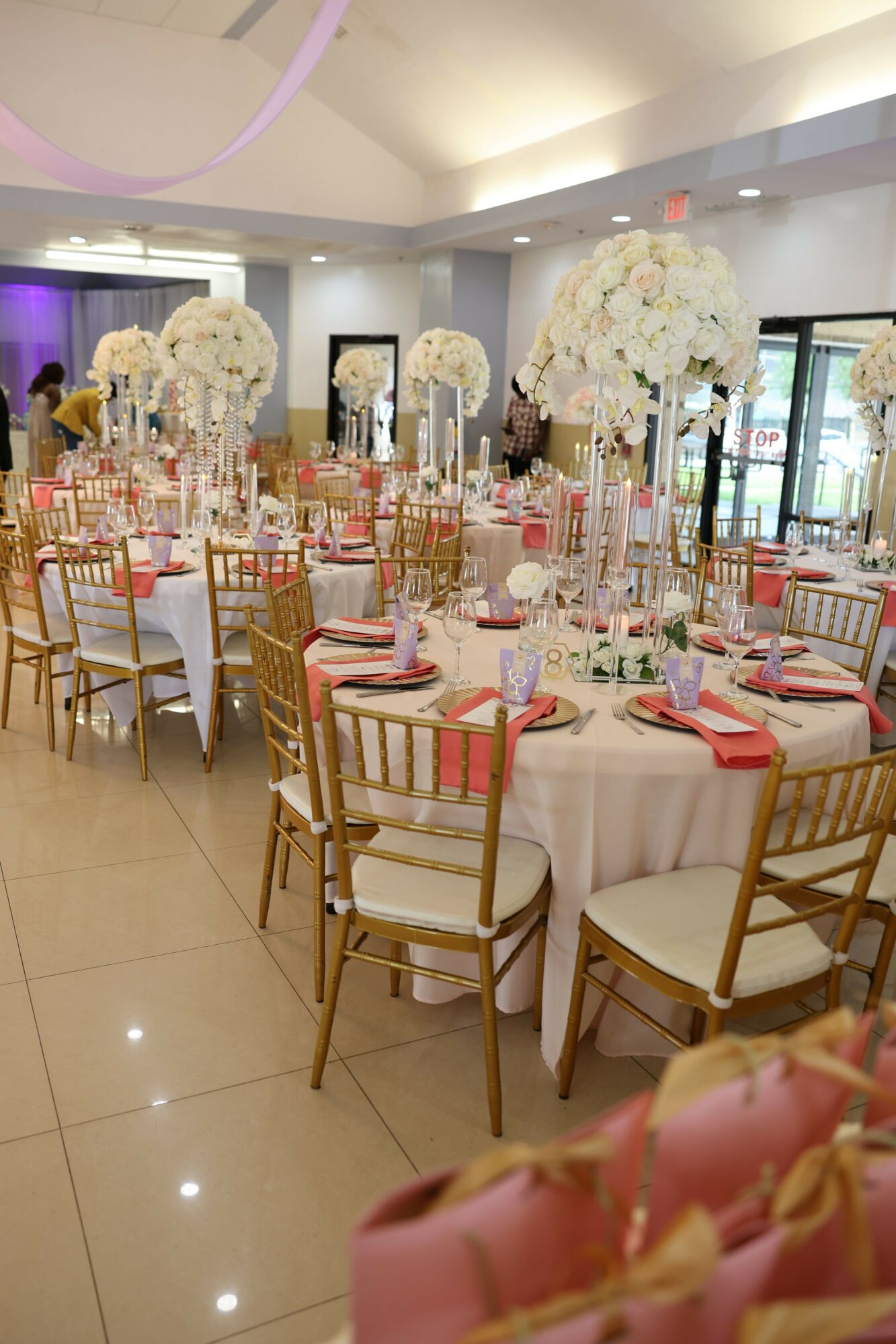 Elegant banquet table set with white tablecloths, pink napkins, floral centerpieces, and gold chairs in a decorated event space.