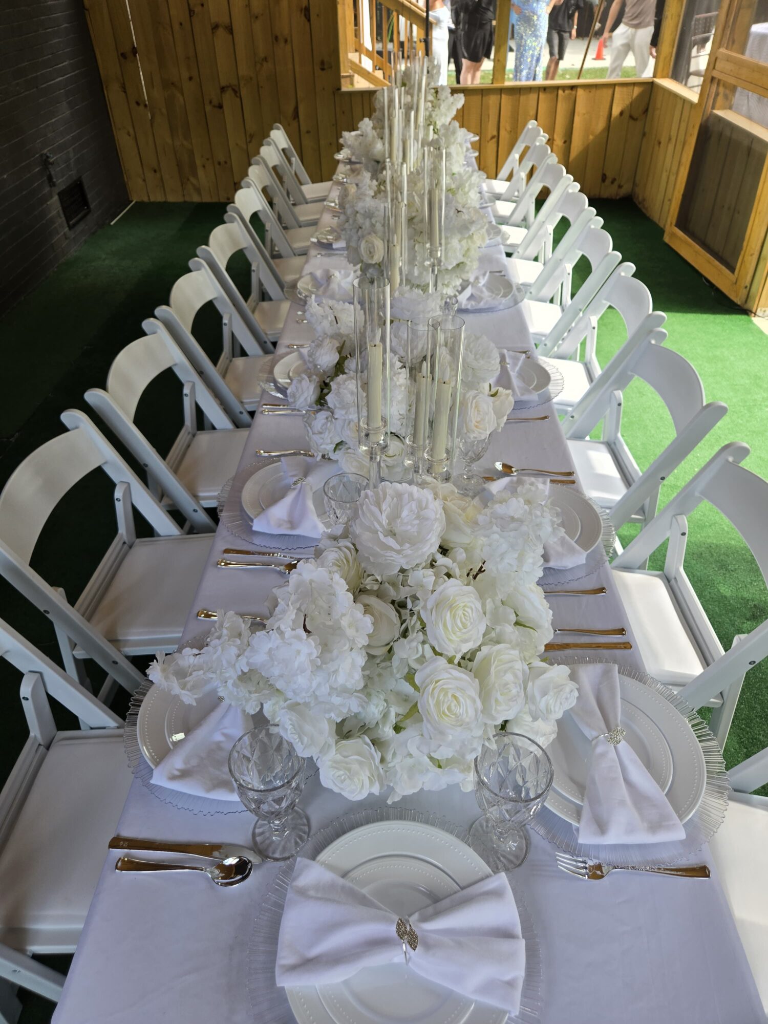 Long table set with white flowers, tall centerpieces, white chairs, and place settings in a wooden room.