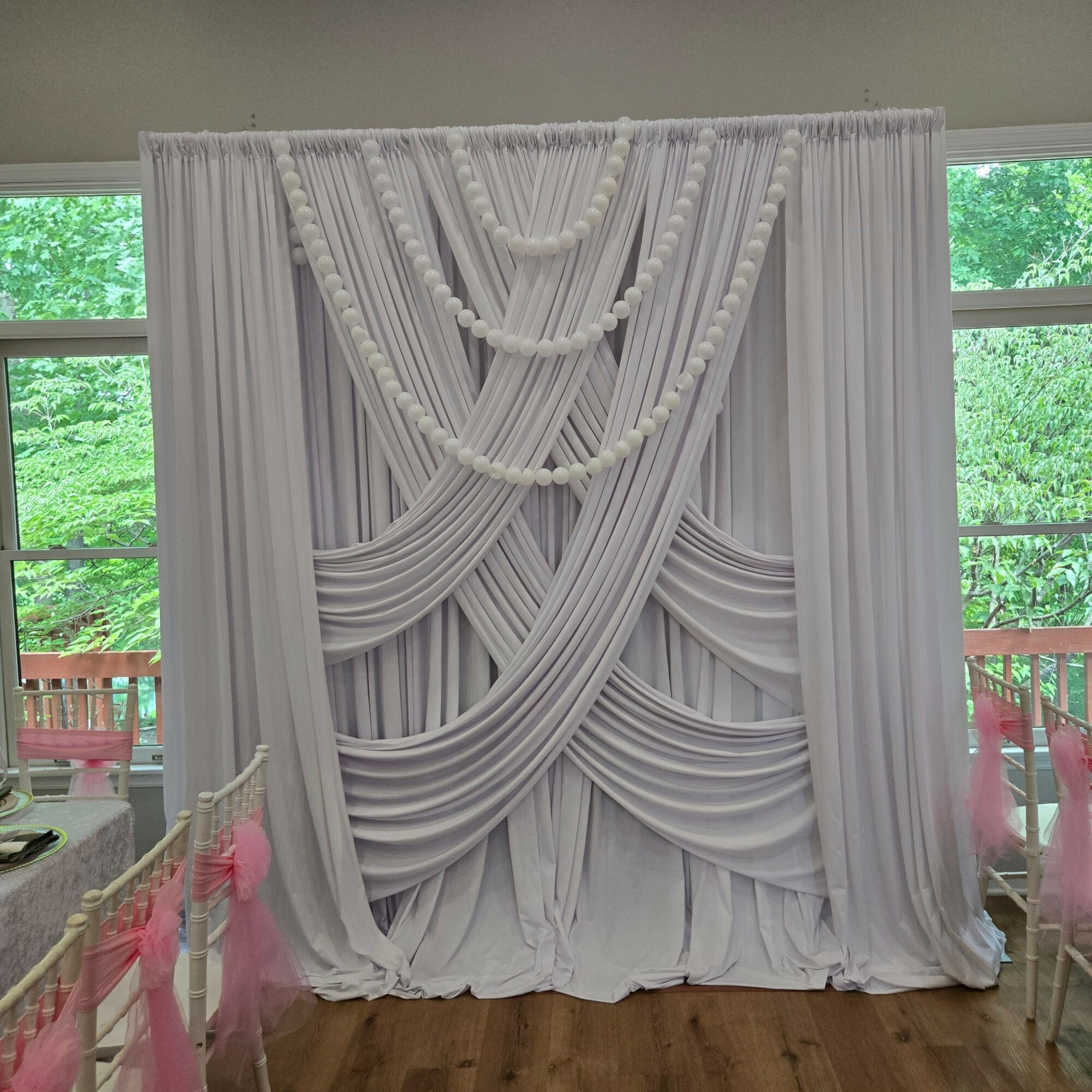 White decorative curtain with intricate draped design hangs in front of windows with green trees outside.