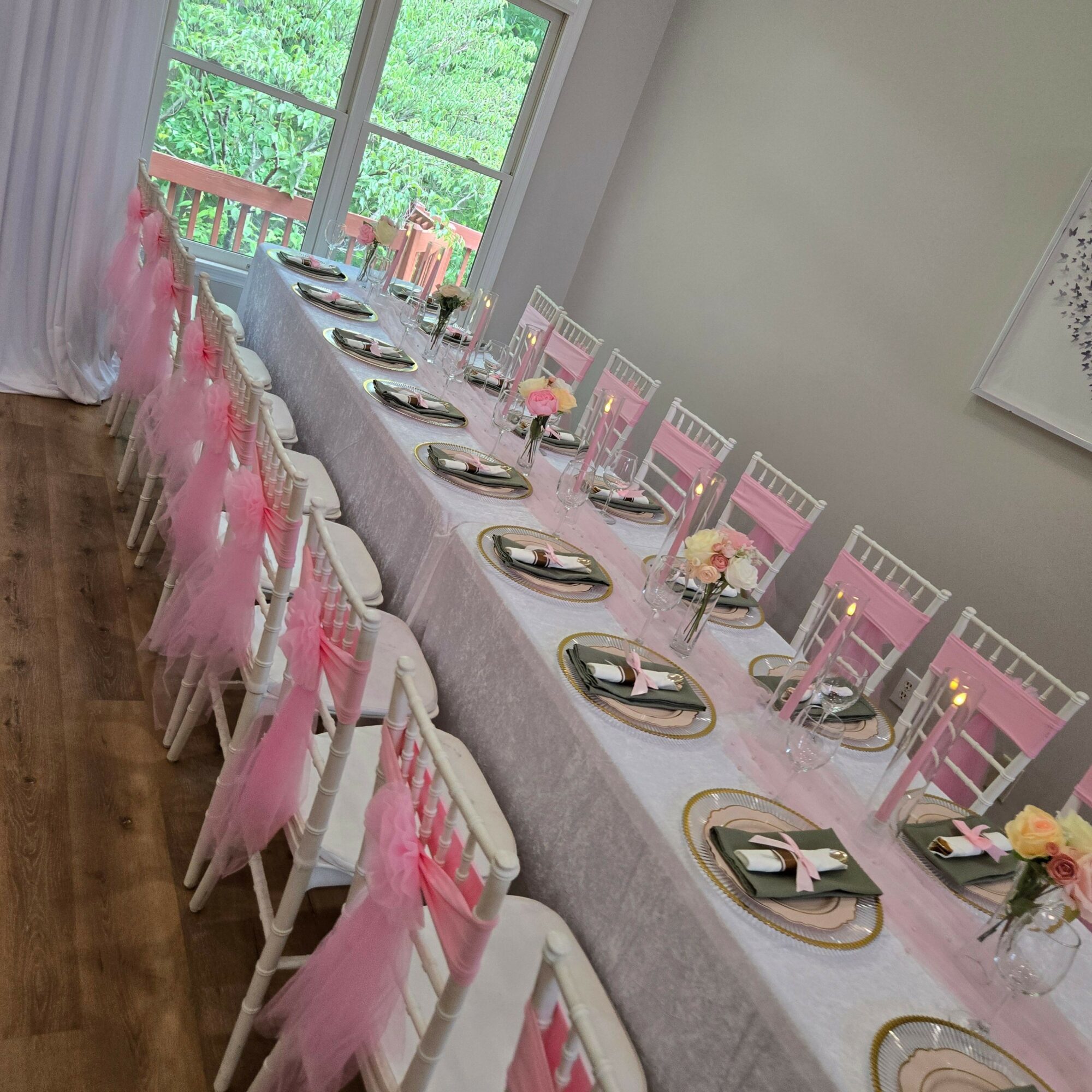 Long table set with pink and gold decorations, plates, glasses, and floral centerpieces, in a bright room with large windows.