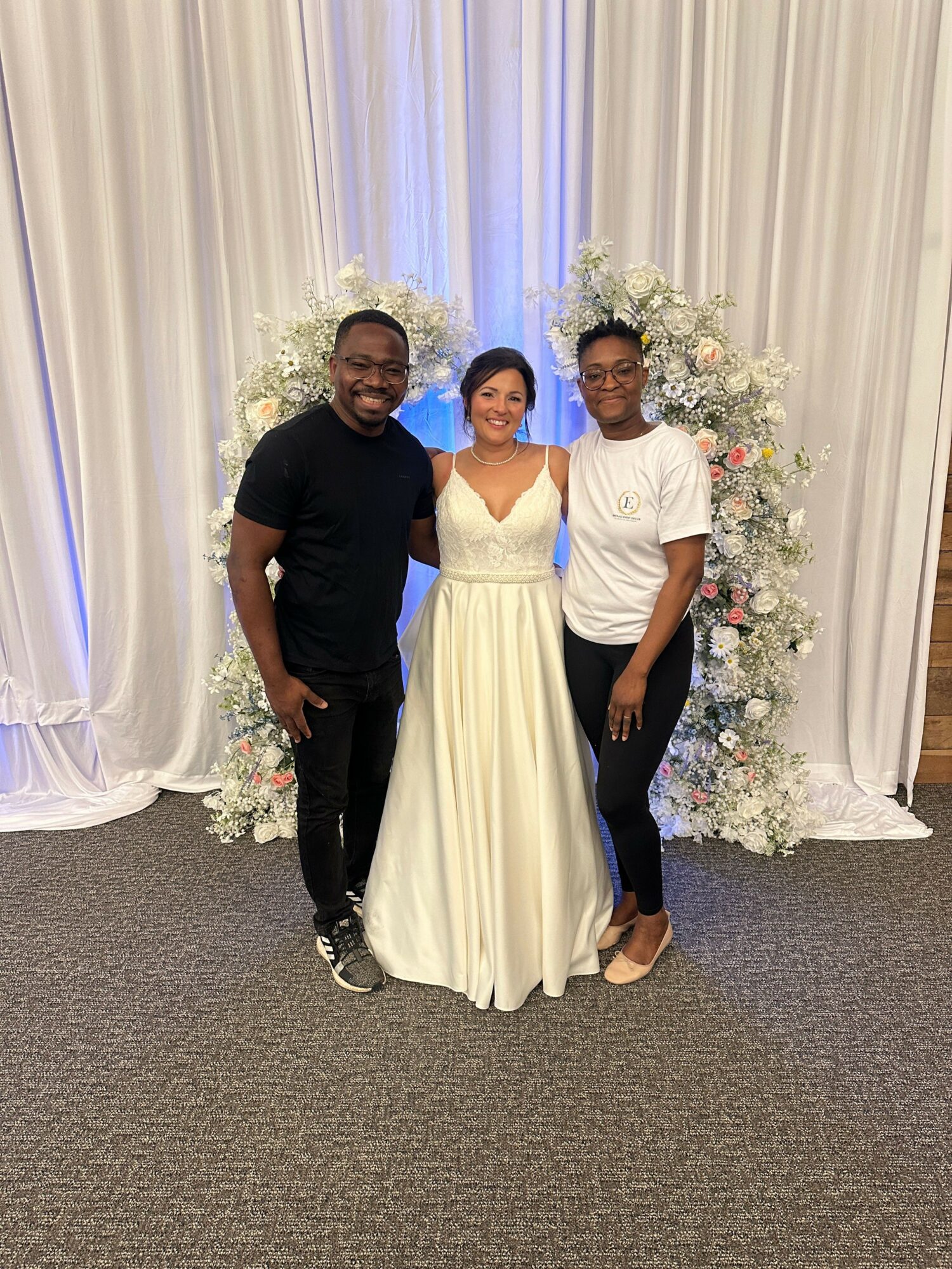 Three people standing in front of floral arrangements and white curtains, smiling, with two women in dresses and a man in a suit.