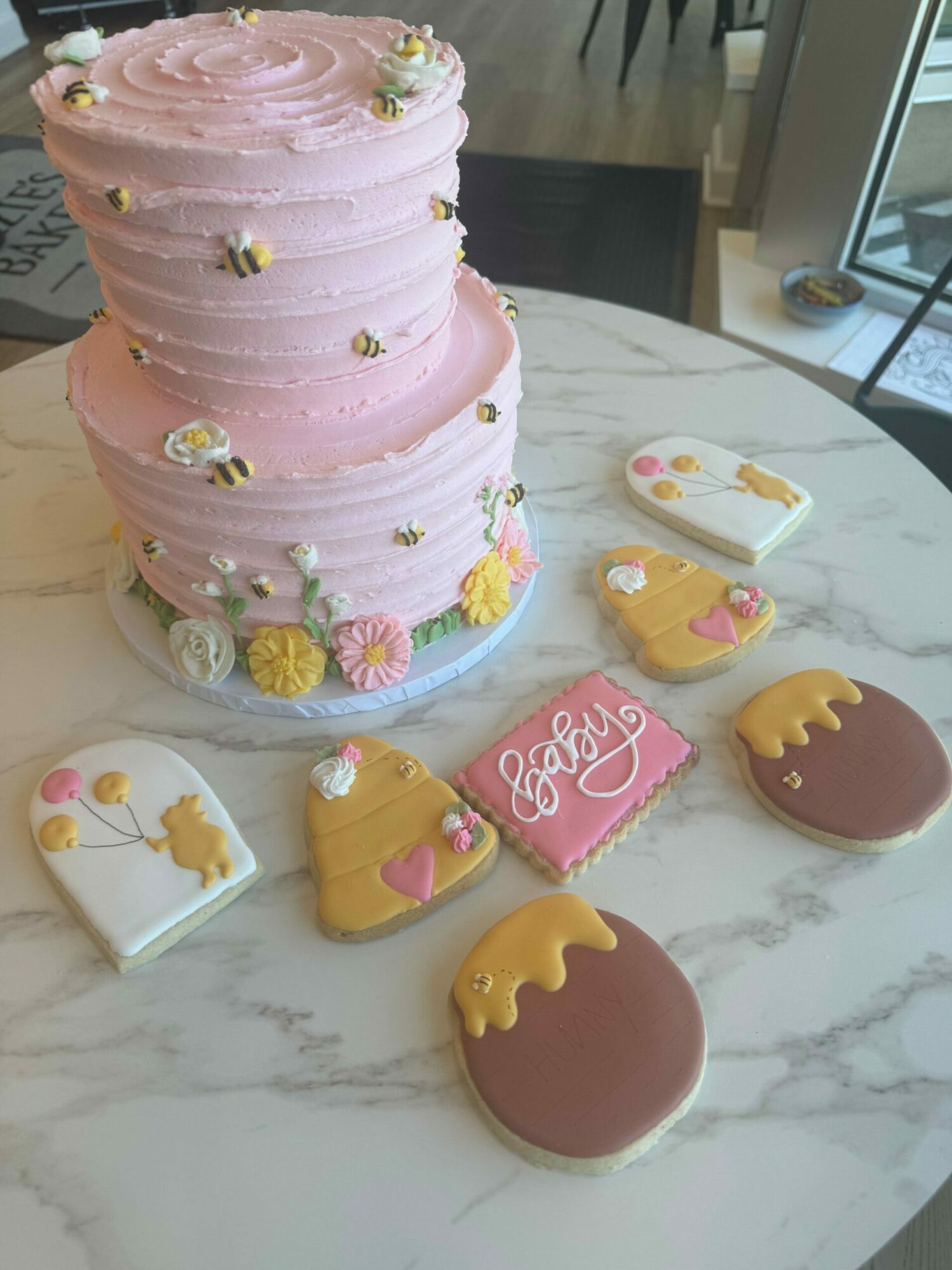 Pink cake with floral decorations and cookies shaped like crowns, ice cream cones, and a cake slice on a marble surface.