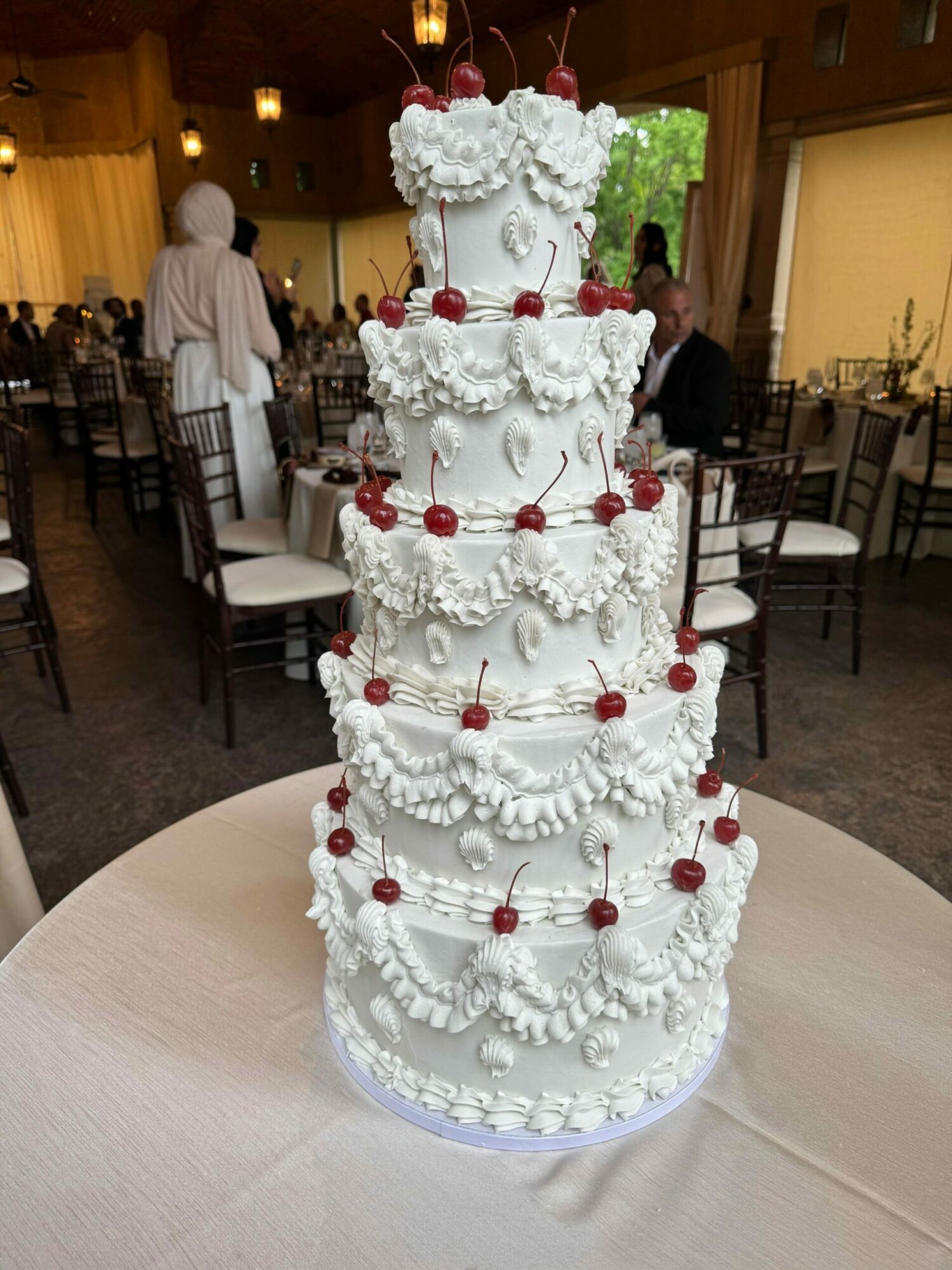 Tall white wedding cake decorated with red cherries and white icing, displayed on a table in a decorated room.