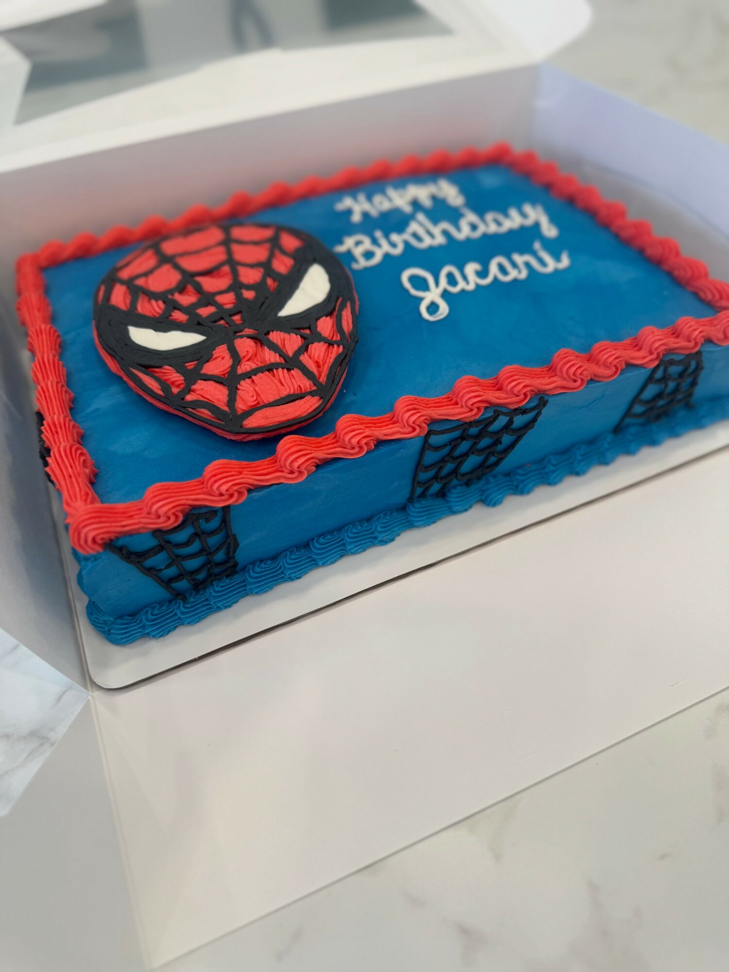 Rectangular birthday cake with blue icing, red piping, Spider-Man face decoration, and a message, in a white box.