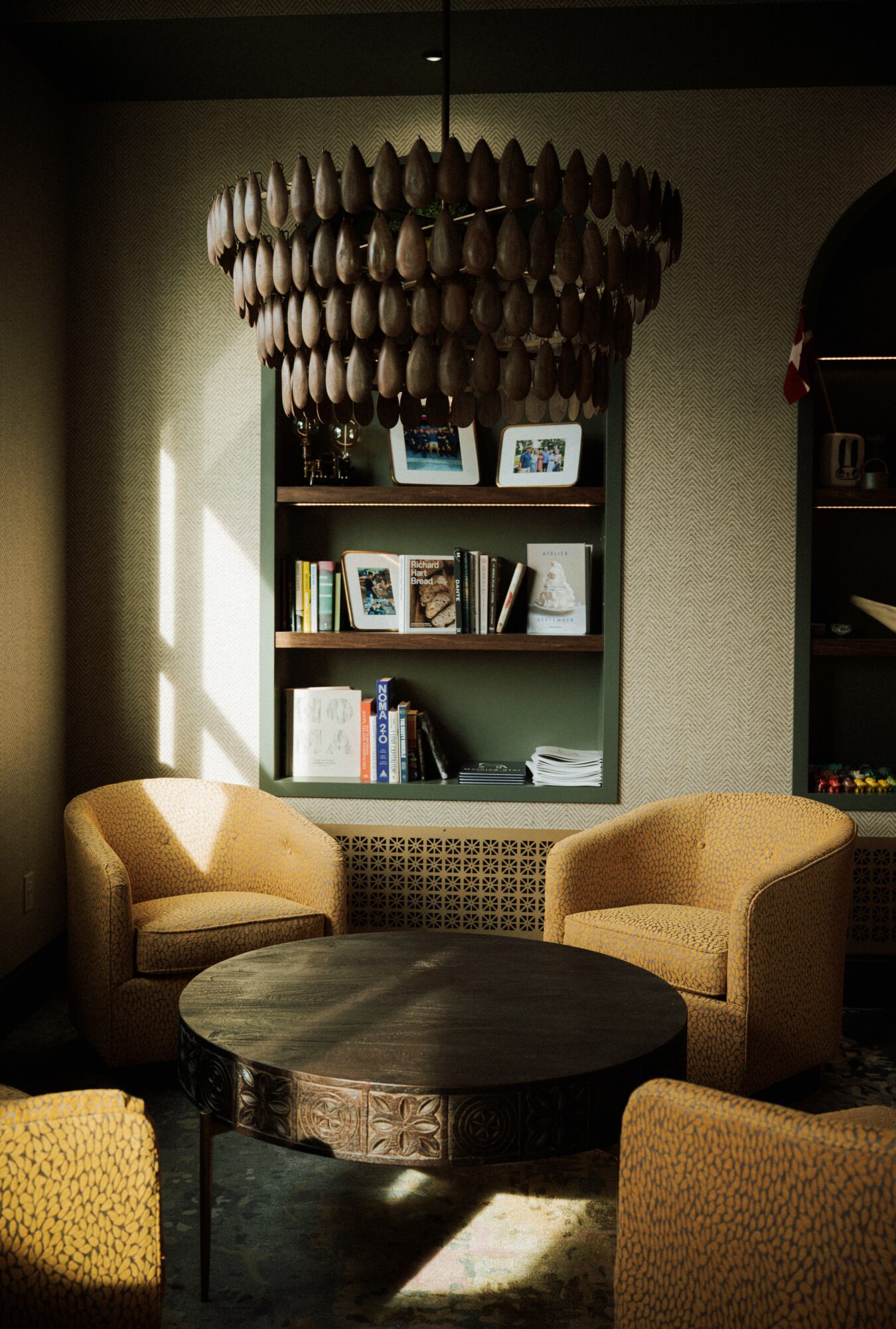 Living room with two beige armchairs around a round wooden table, bookshelf with books and photos, chandelier overhead.