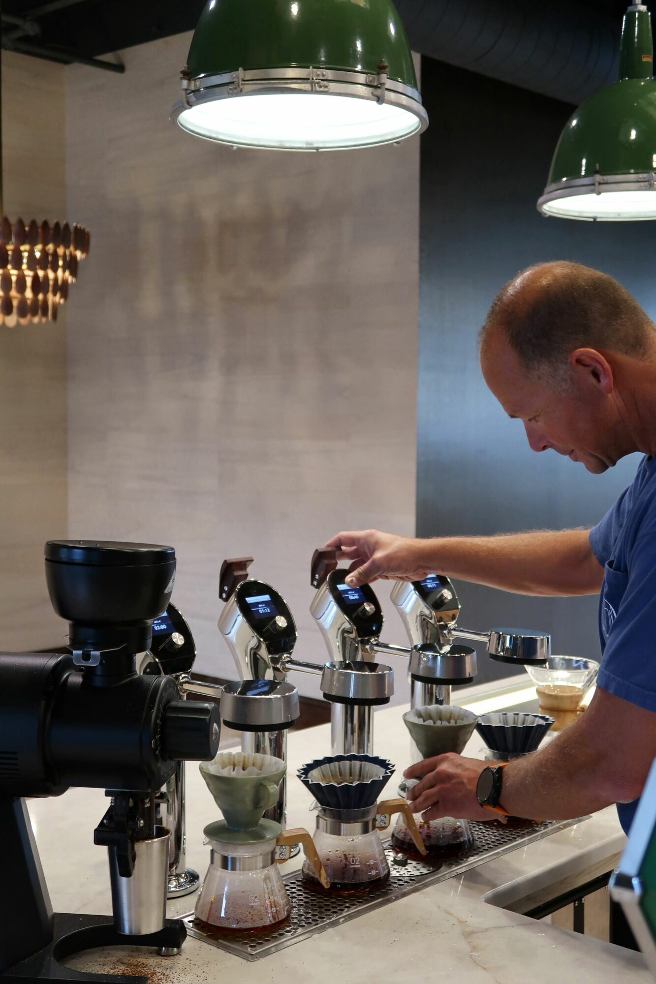 Person pouring coffee into cups on a table with a camera in foreground, two green lamps overhead, and a metallic wall background.