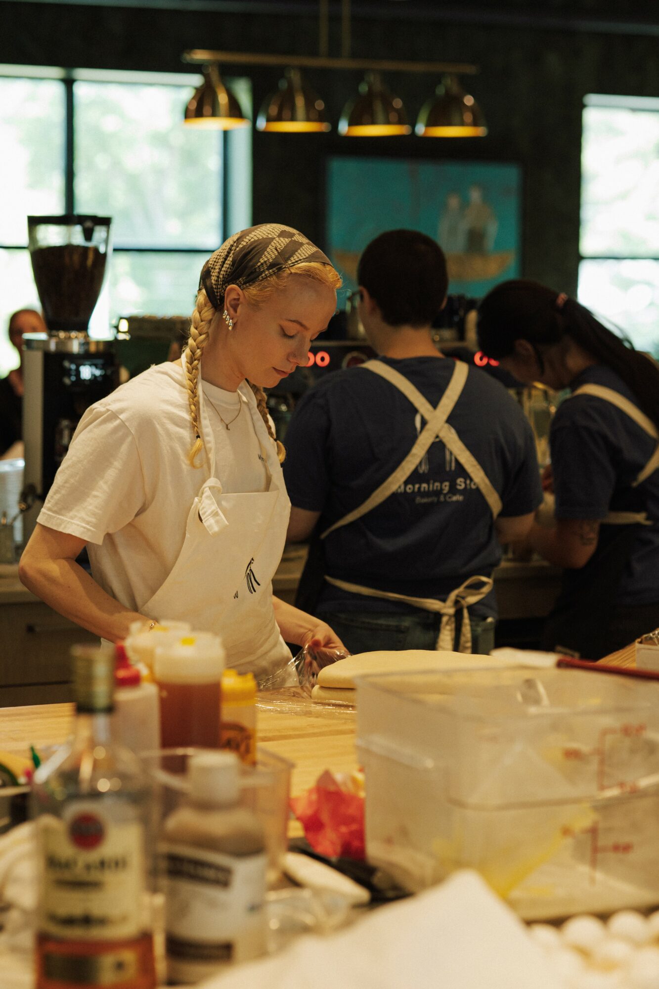 Young woman with braided hair working in a kitchen with other people, ingredients and kitchen tools visible.