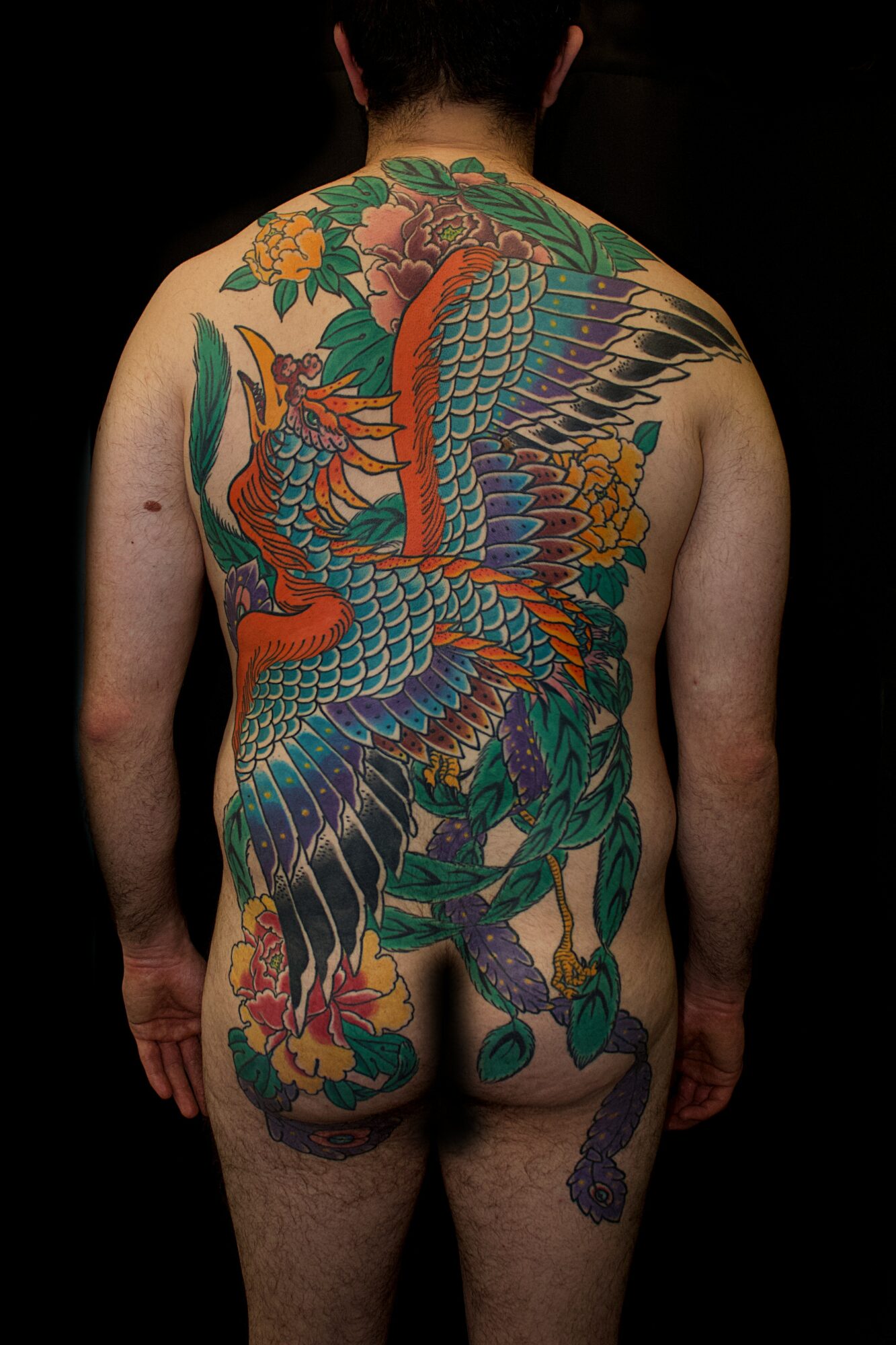Back view of a person with a large, colorful dragon tattoo covering the entire back and extending to the legs.
