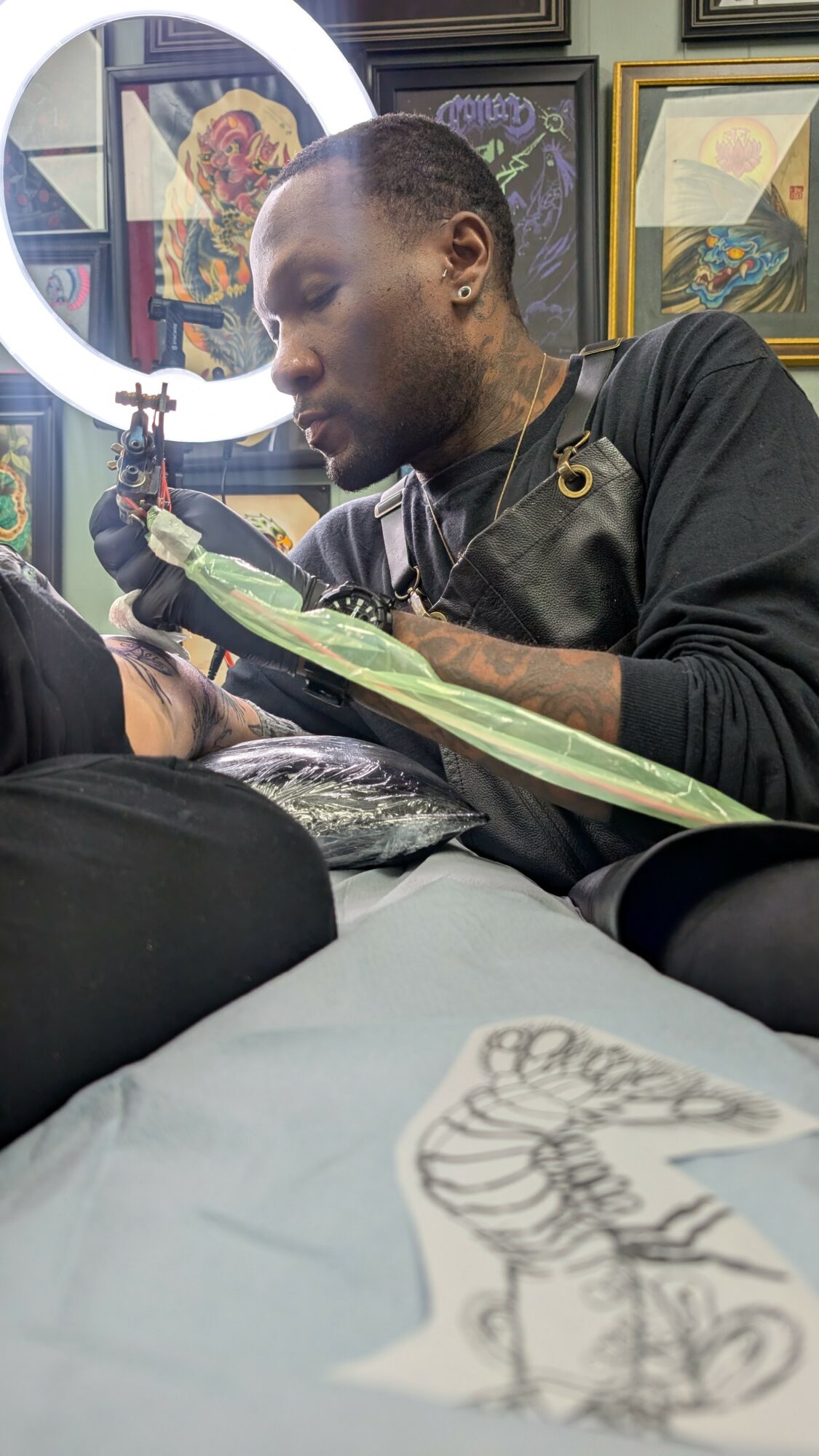 Tattoo artist working on a client's arm in a tattoo studio with framed artwork on the wall.