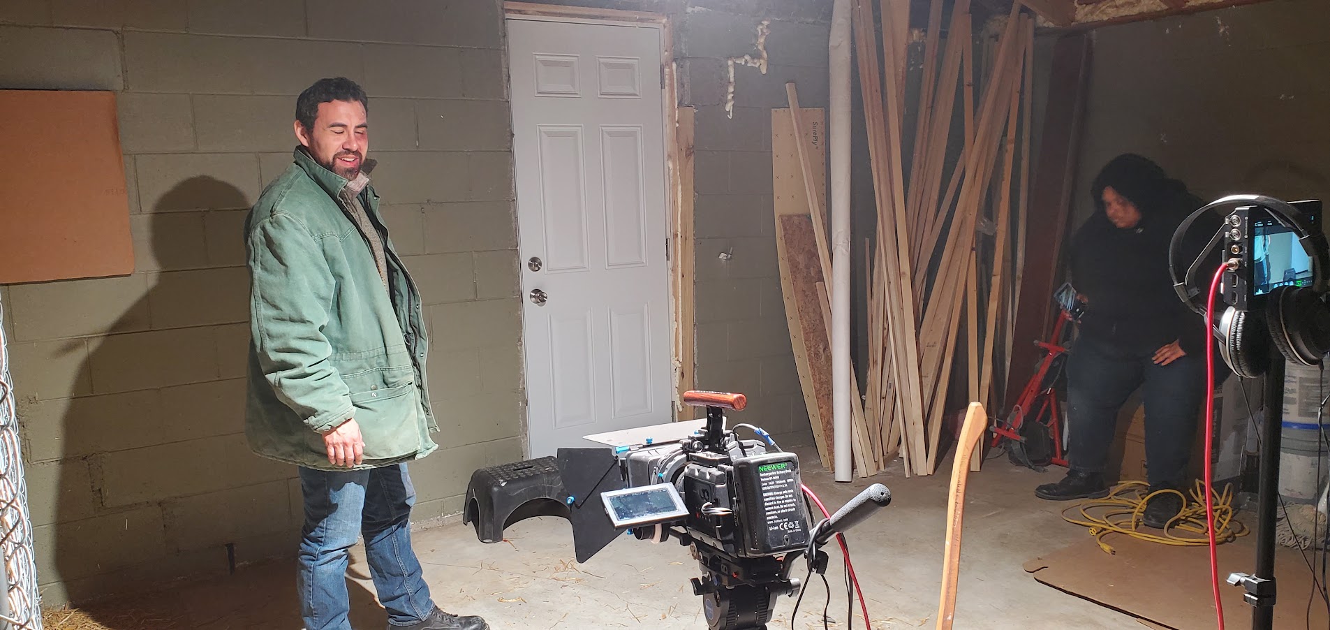 Man with beard in green jacket standing in a workshop with tools and wood panels, smiling at the camera.