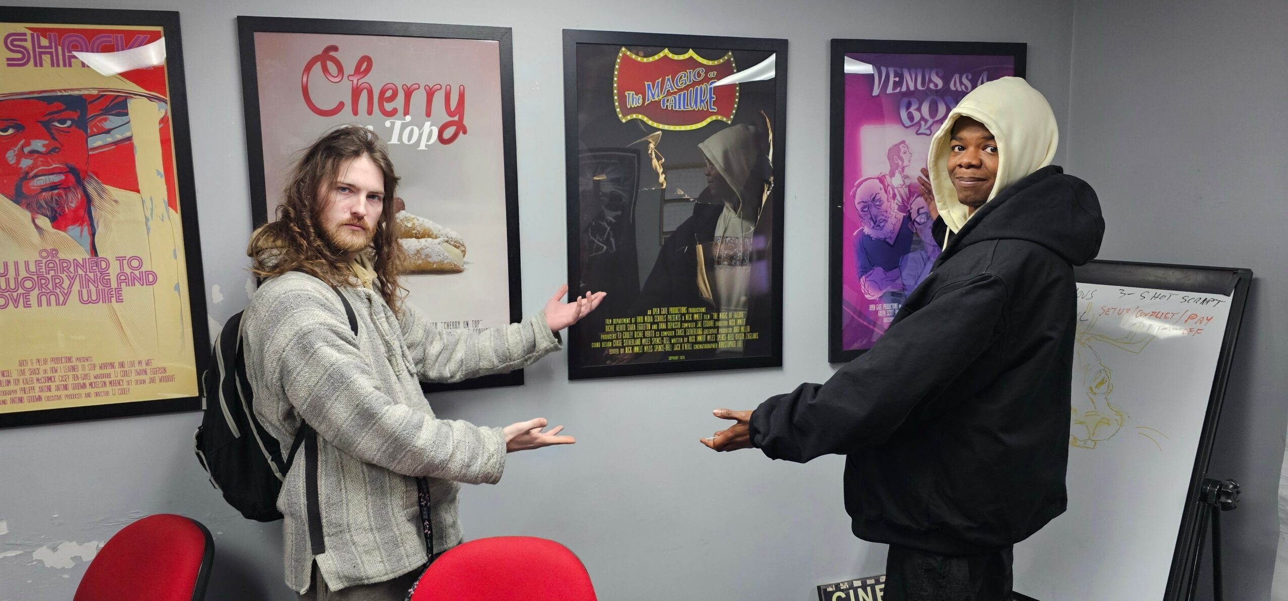 Two people standing in front of a wall with framed posters, pointing at the posters, in a room with gray walls.
