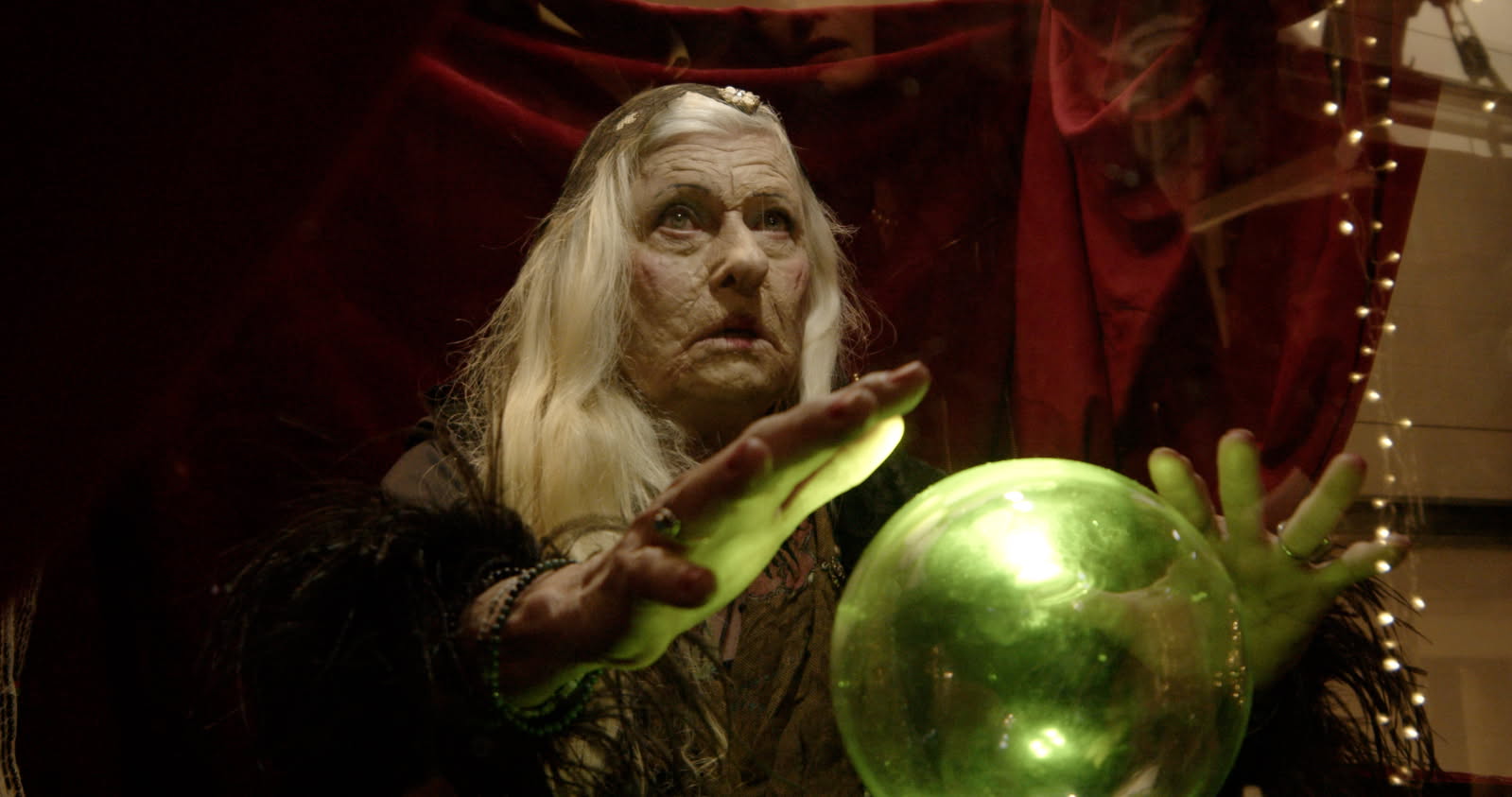 An elderly woman with long gray hair reaches toward a glowing green orb with both hands.