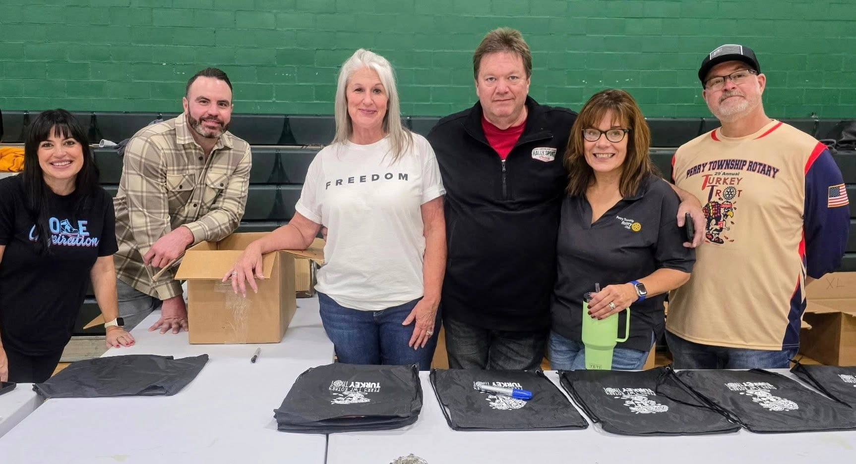 Group of six people standing behind a table with black bags, smiling, in front of a green wall.