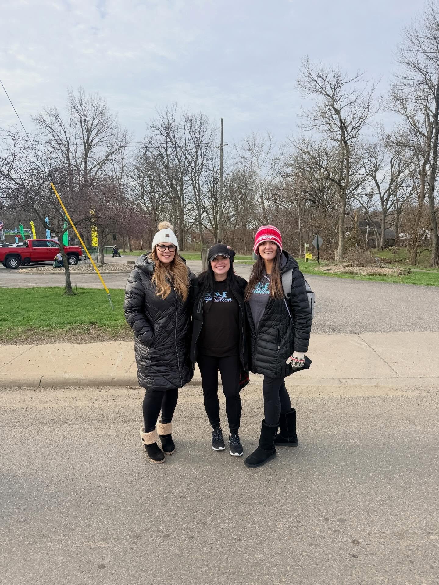 Three women standing outdoors on a paved area, wearing winter coats and hats, with trees and a playground in the background.