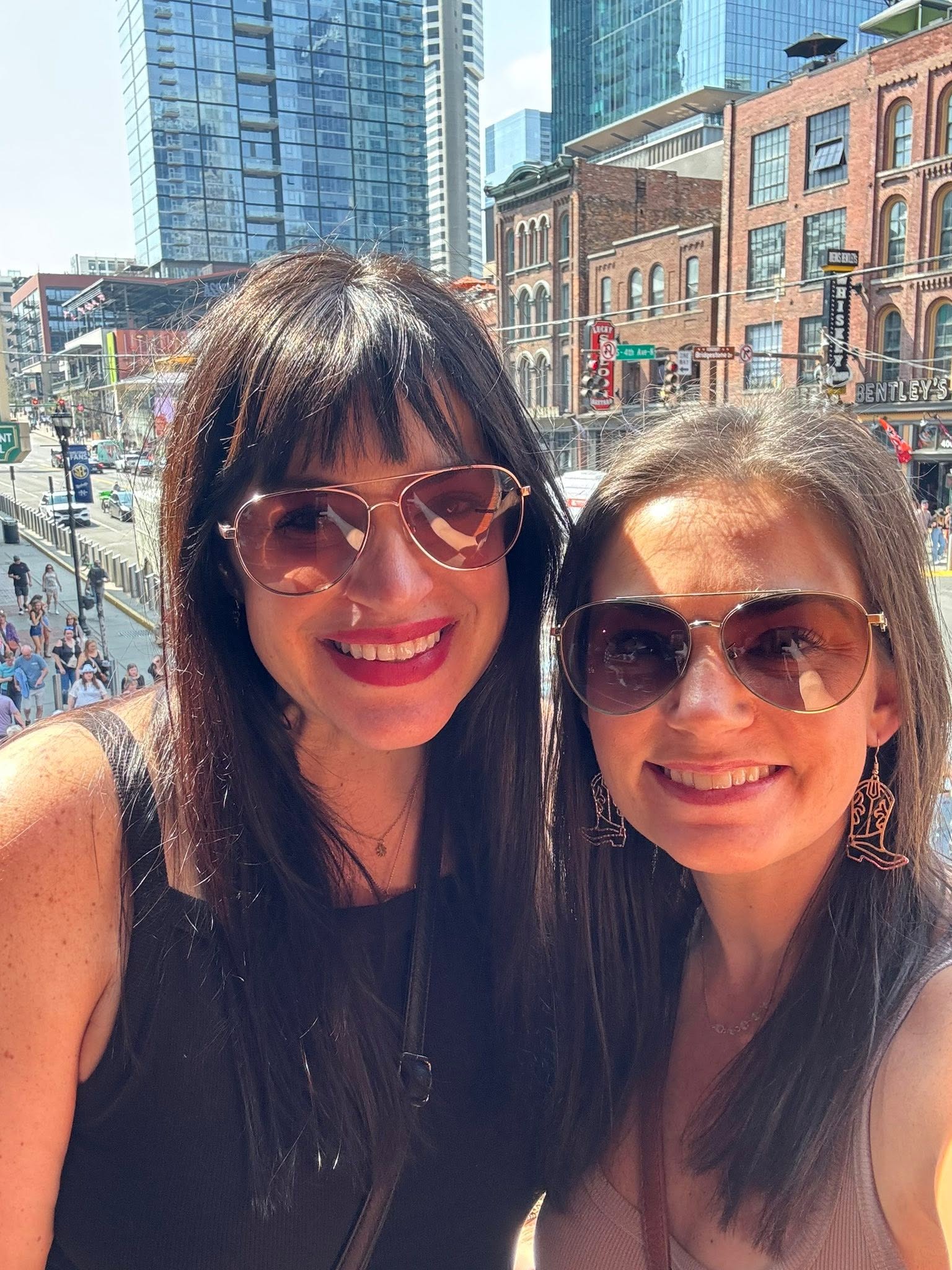 Two women smiling outdoors with city buildings and streets in the background, both wearing sunglasses and dark clothing.