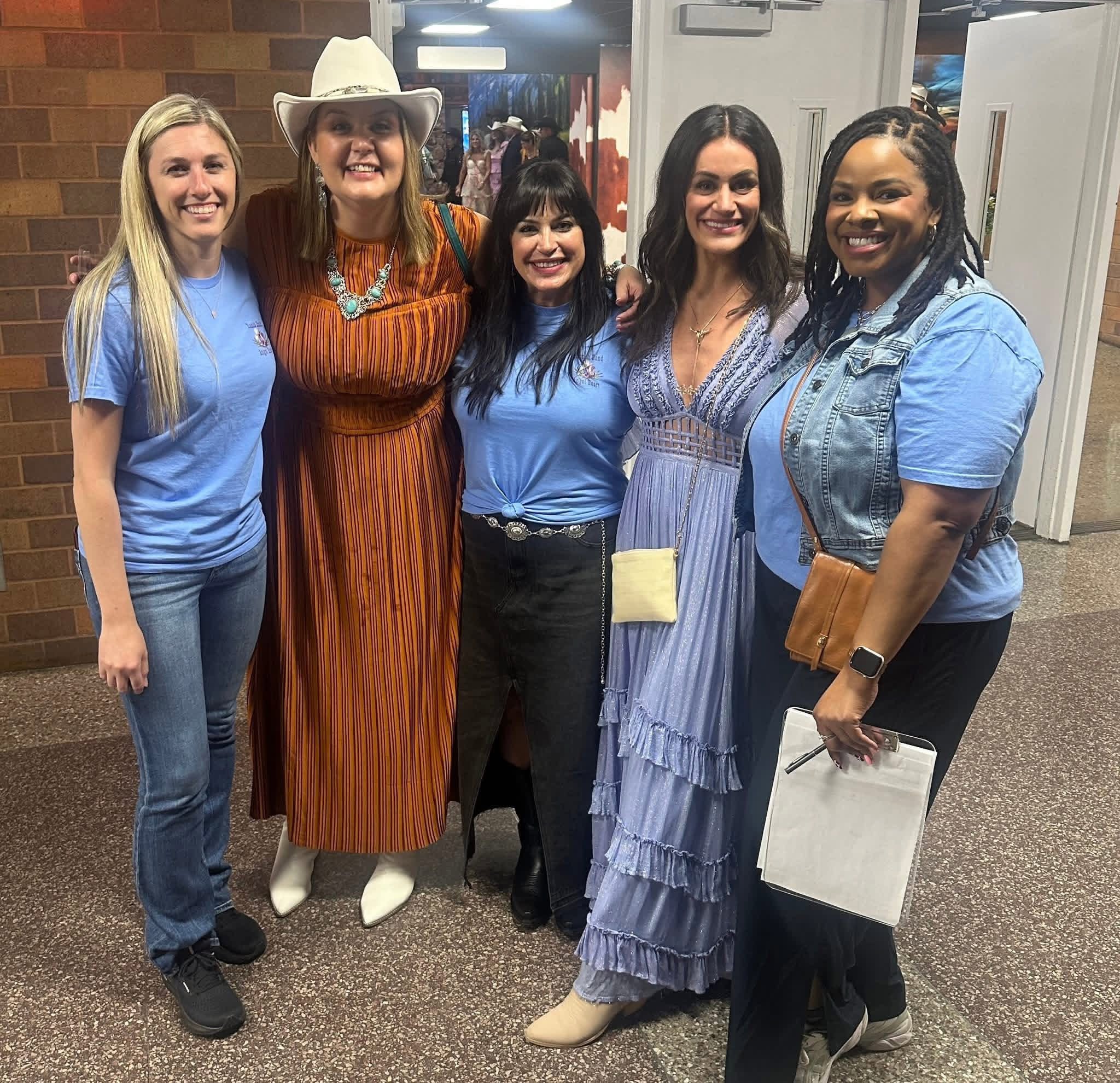 Five women standing together indoors, smiling, with casual and dressy clothing, some holding papers or folders.