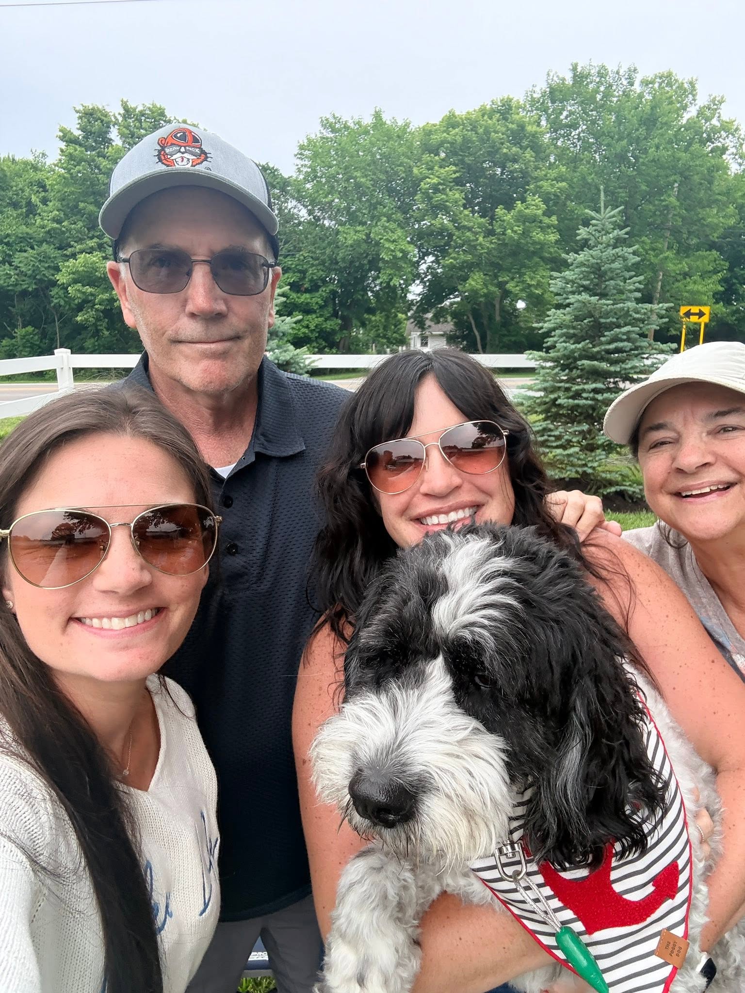 Four people and a dog outdoors with trees in the background, smiling and wearing sunglasses.