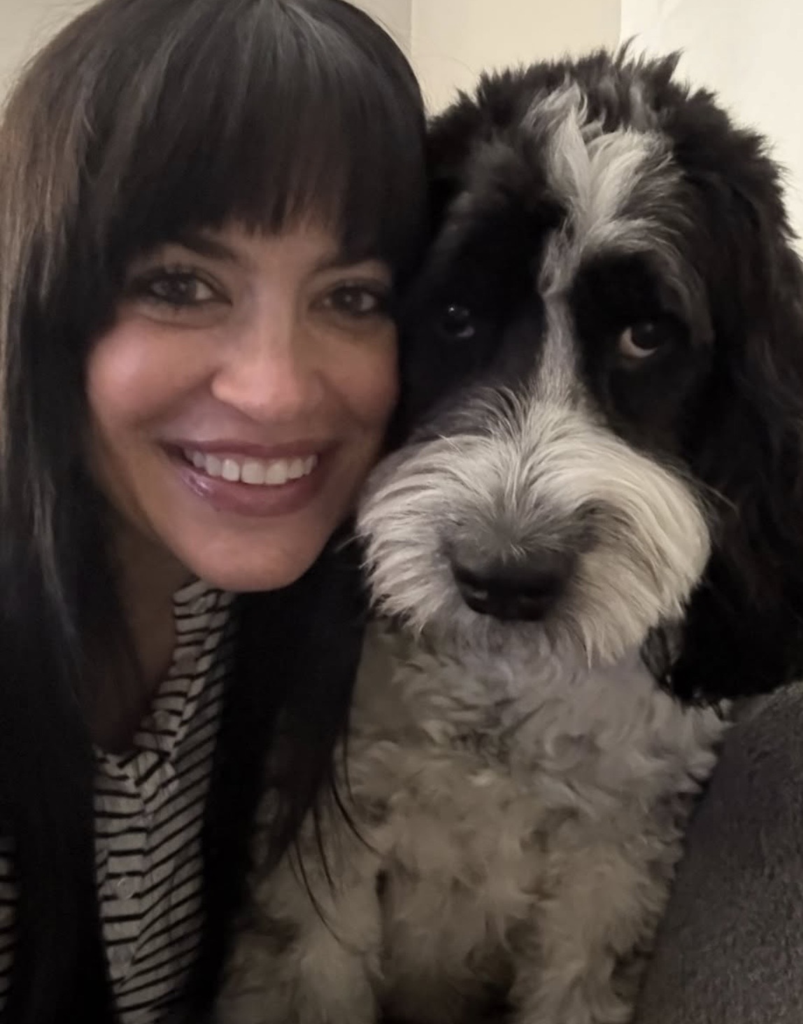 Woman smiling with a black and white fluffy dog close to her face, both looking at the camera.