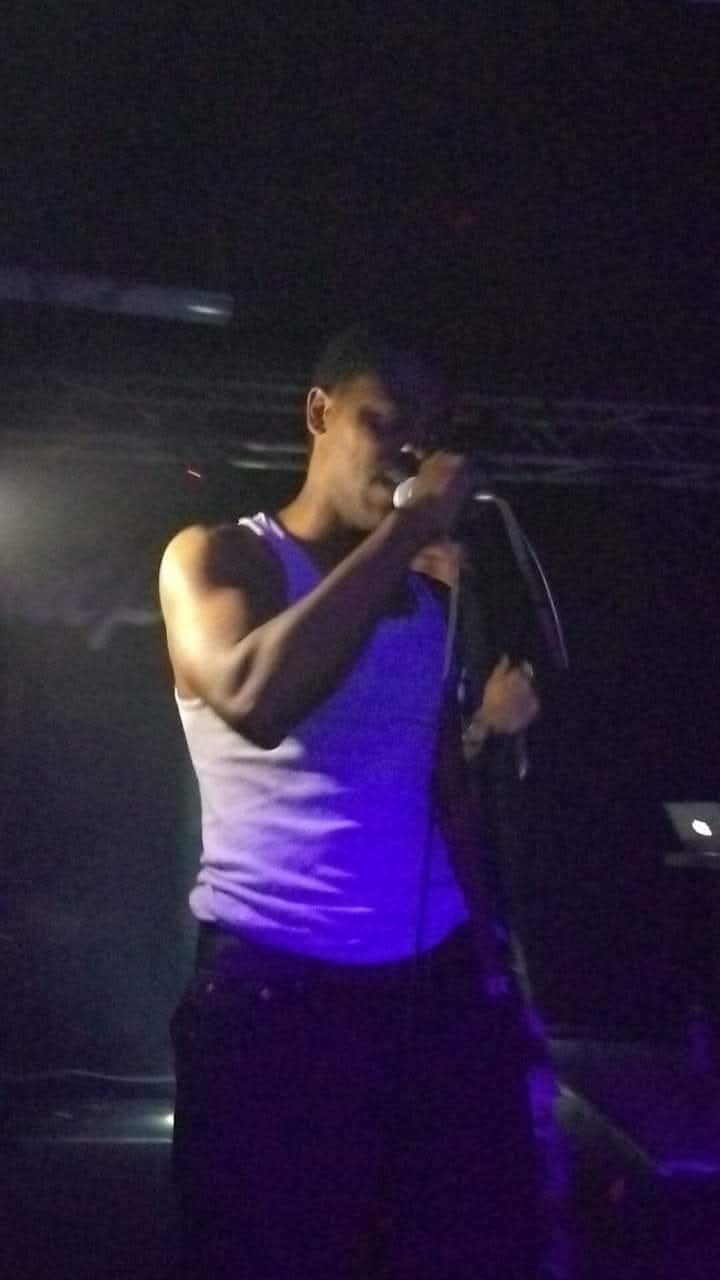 Person singing into a microphone on stage, wearing a sleeveless top, with dark background and stage lighting.