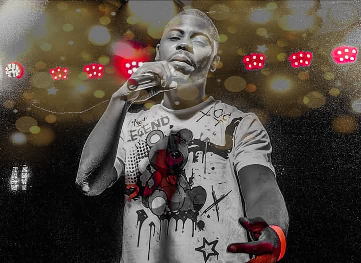 Person singing into a microphone with a red plush toy in front, stage lights in background, wearing a graphic t-shirt.