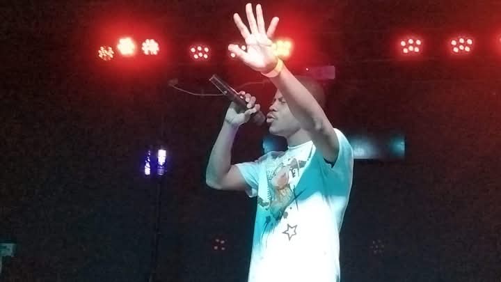 Person on stage holding microphone, raising hand, with stage lights above, wearing a light-colored shirt with star design.