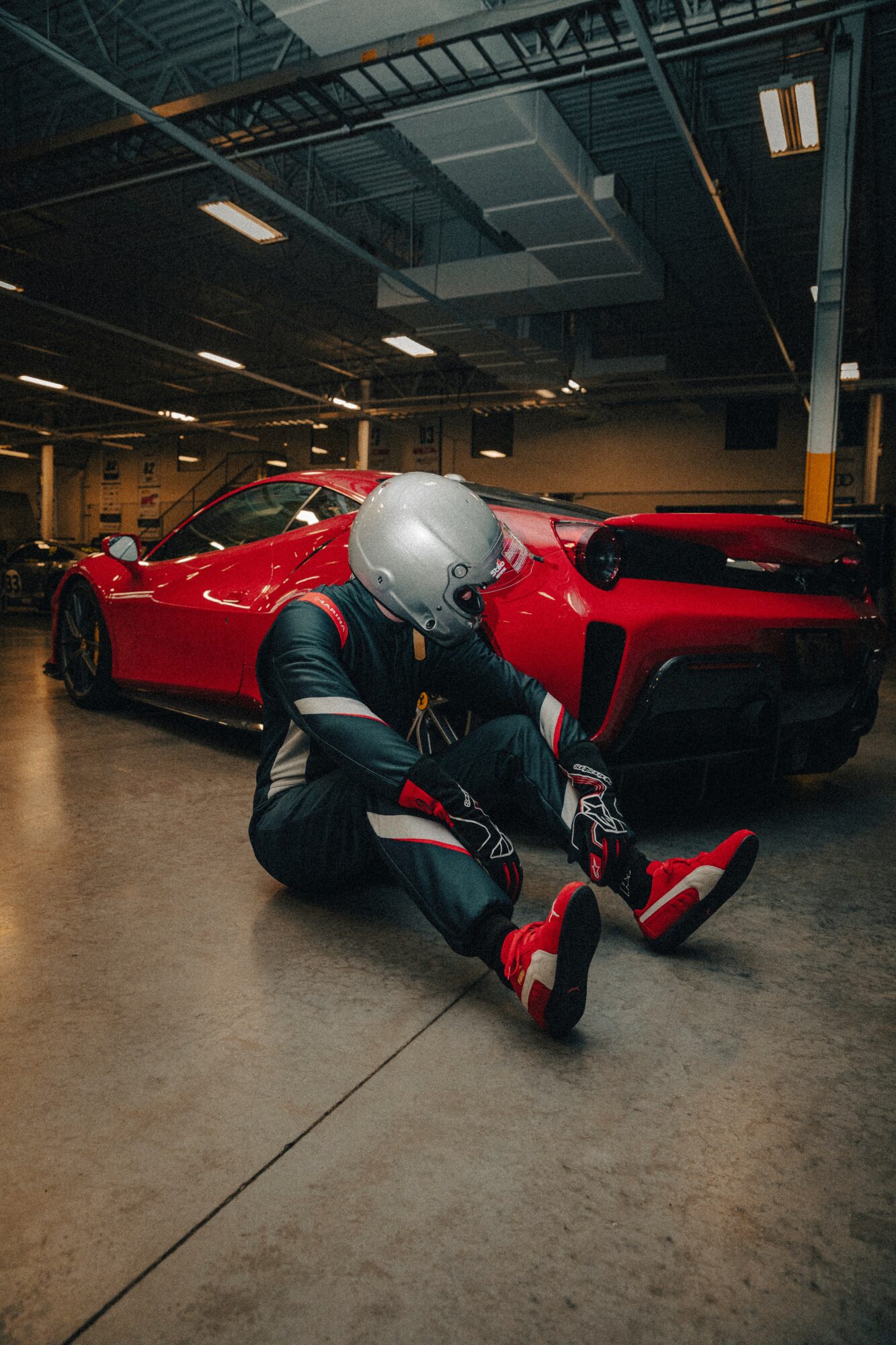 Person in racing suit and helmet sitting on the floor next to a red sports car in an indoor garage.