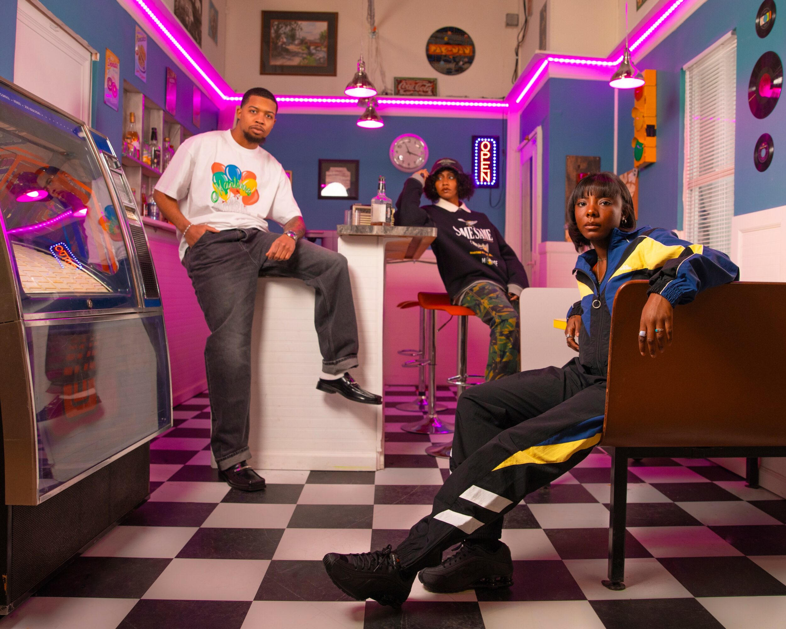 Three people in a colorful retro-style diner with checkered floor, neon lights, and vintage decor, posing casually.