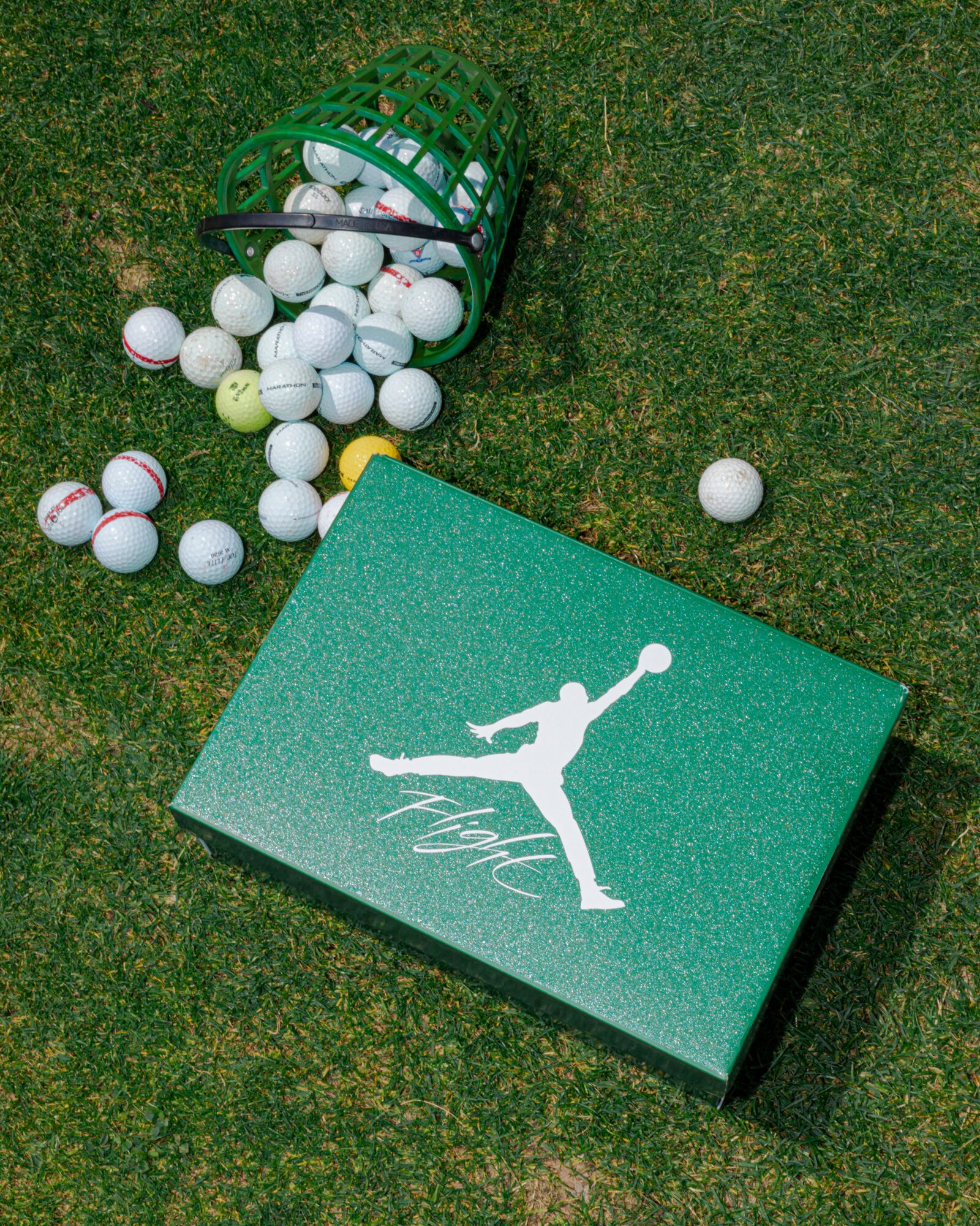 Green box with a white figure of a person playing tennis, on grass with scattered golf balls and a green basket.