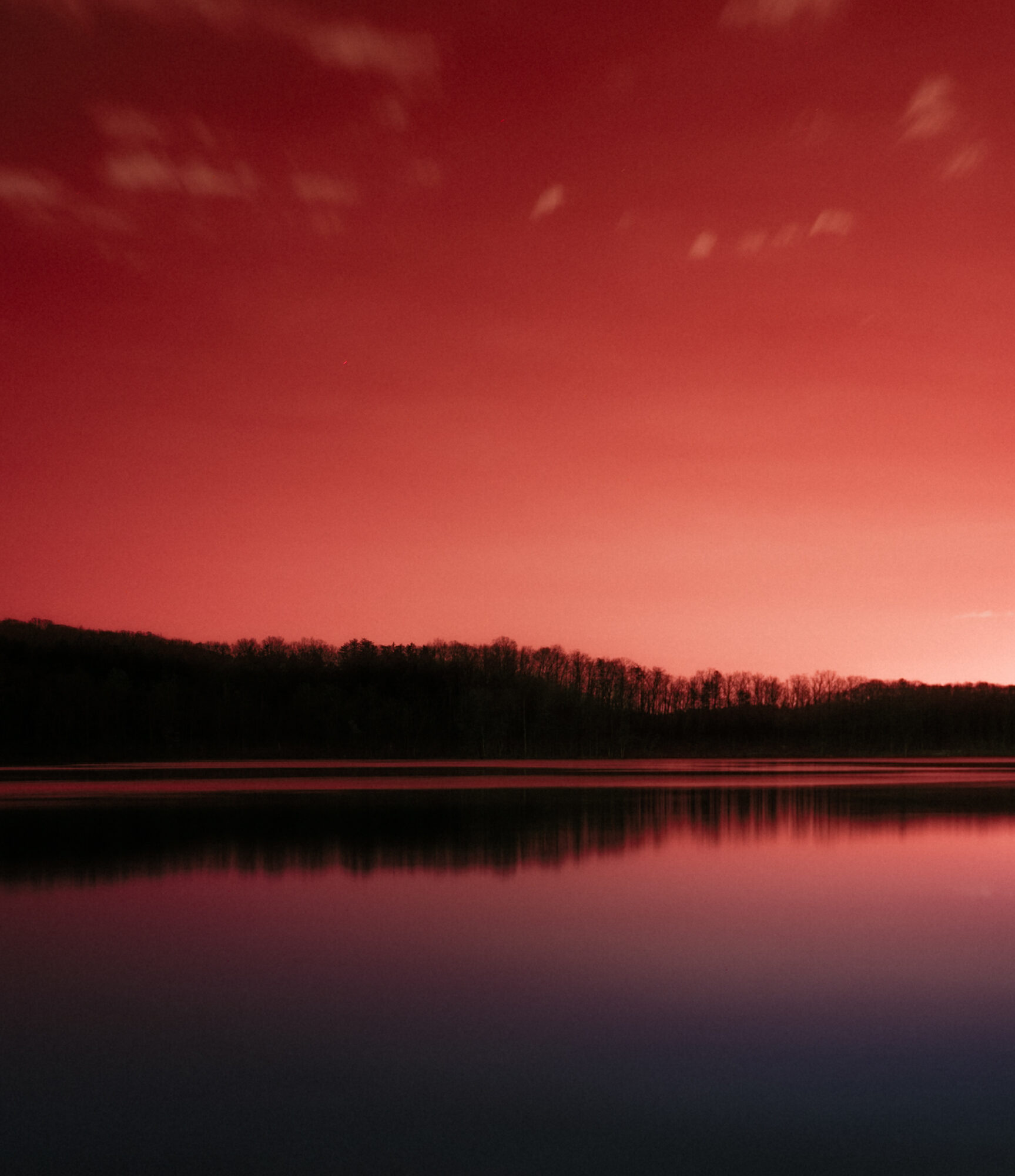 Nighttime landscape with a pinkish-red sky, a dark treeline, and a calm body of water reflecting the sky.